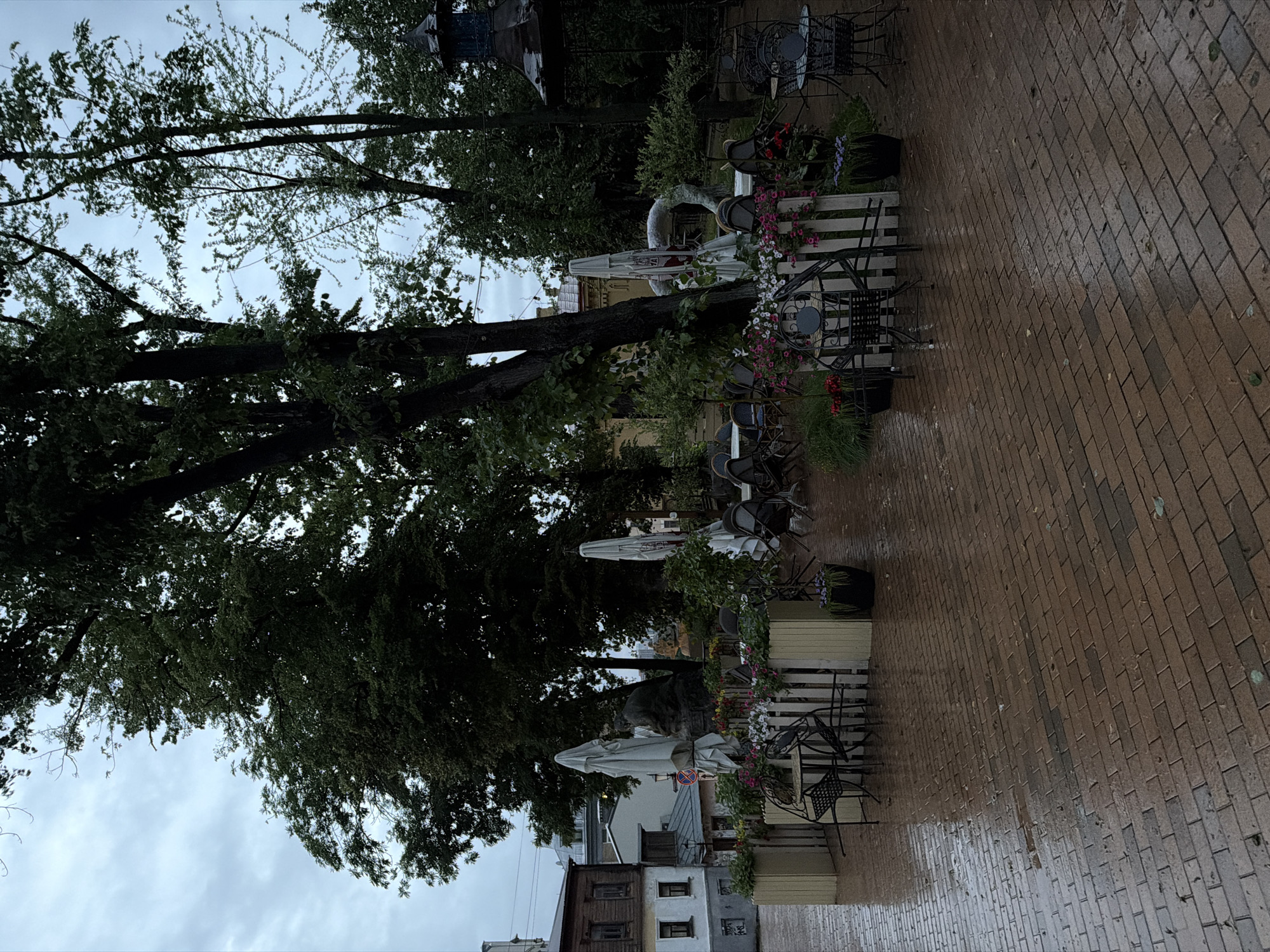 The image depicts an outdoor cafe area with red brick paving stones, wet from recent rain, suggesting an overcast or inclement weather day. Several tables and chairs, some made of dark metal, are arranged on the paved area. White umbrellas are partially furled over some tables. Planters with flowers and greenery are placed around the seating area, interspersed with white picket fencing. In the background, there are trees and buildings, including a distinctive wooden-clad structure on the left. To the right, a dark gazebo-like structure with decorative elements is visible amongst the trees. A statue of a person is positioned behind a cluster of umbrellas. The overall atmosphere is subdued and quiet, possibly due to the weather.