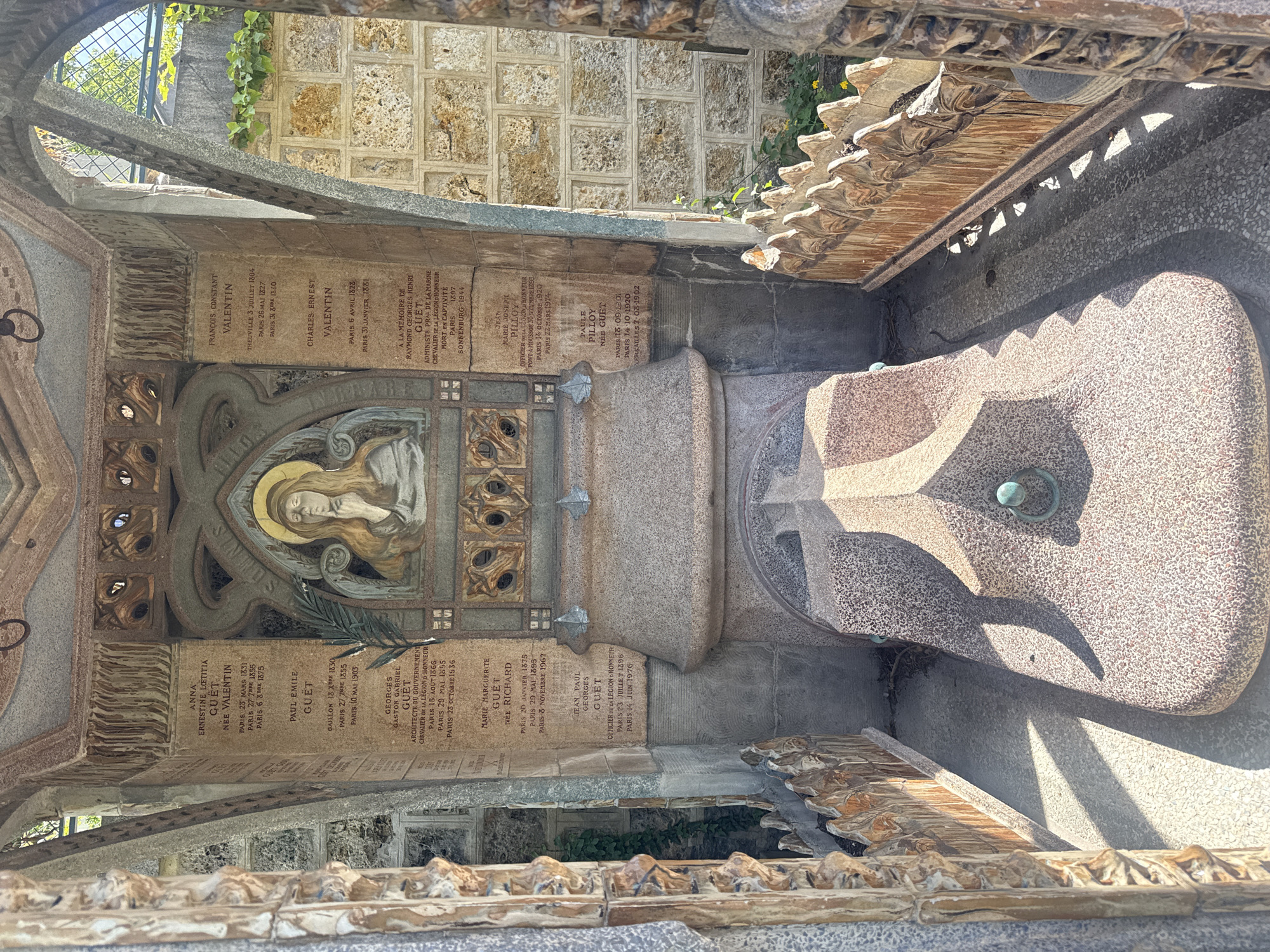 The image shows an above-eye-level view of a tomb or mausoleum likely located in a cemetery. The structure is elaborately decorated with carvings, inscriptions, and a central bas-relief depicting a person (possibly a saint) with a halo. There are inscriptions on the left and right walls, bearing the names "GUET" and "VALENTIN" along with birth and death dates in Paris, France, and elsewhere. A stone sarcophagus or basin is in the center, suggesting a place of rest or commemoration. The architectural style is ornate and possibly dates to the late 19th or early 20th century. The presence of stone blocks in the background suggests this is an outdoor setting within a walled cemetery area. The location is Paris, France.