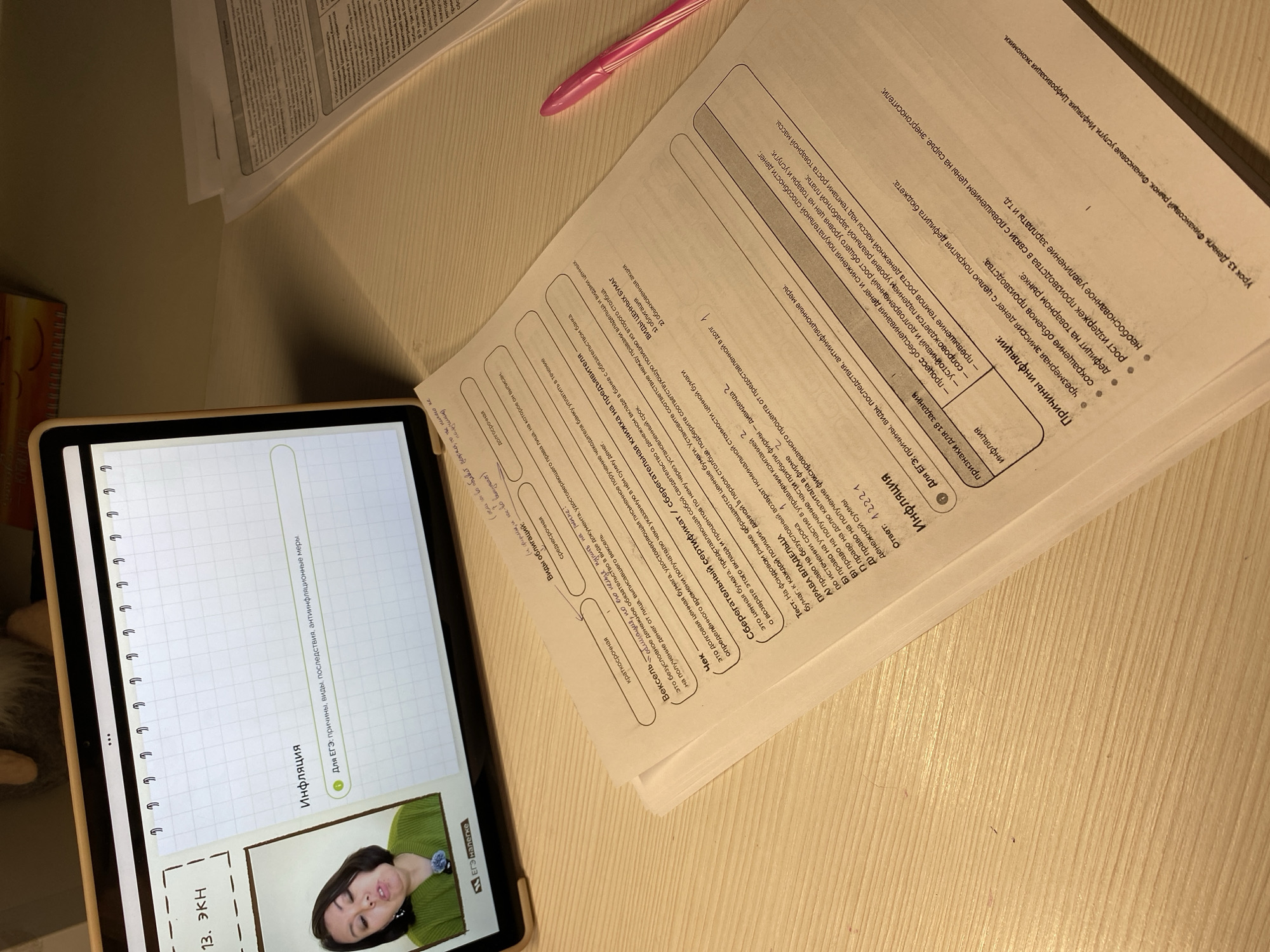 A desk with a tablet, some papers, and a pen on it. The tablet has a study session on screen, with the title "Inflation". The papers on the desk also seem to be economics related, with Russian text on them. A pink pen lies on top of the papers. The overall environment looks like a study space. There's no indication of location such as Votkinsk, Russia.