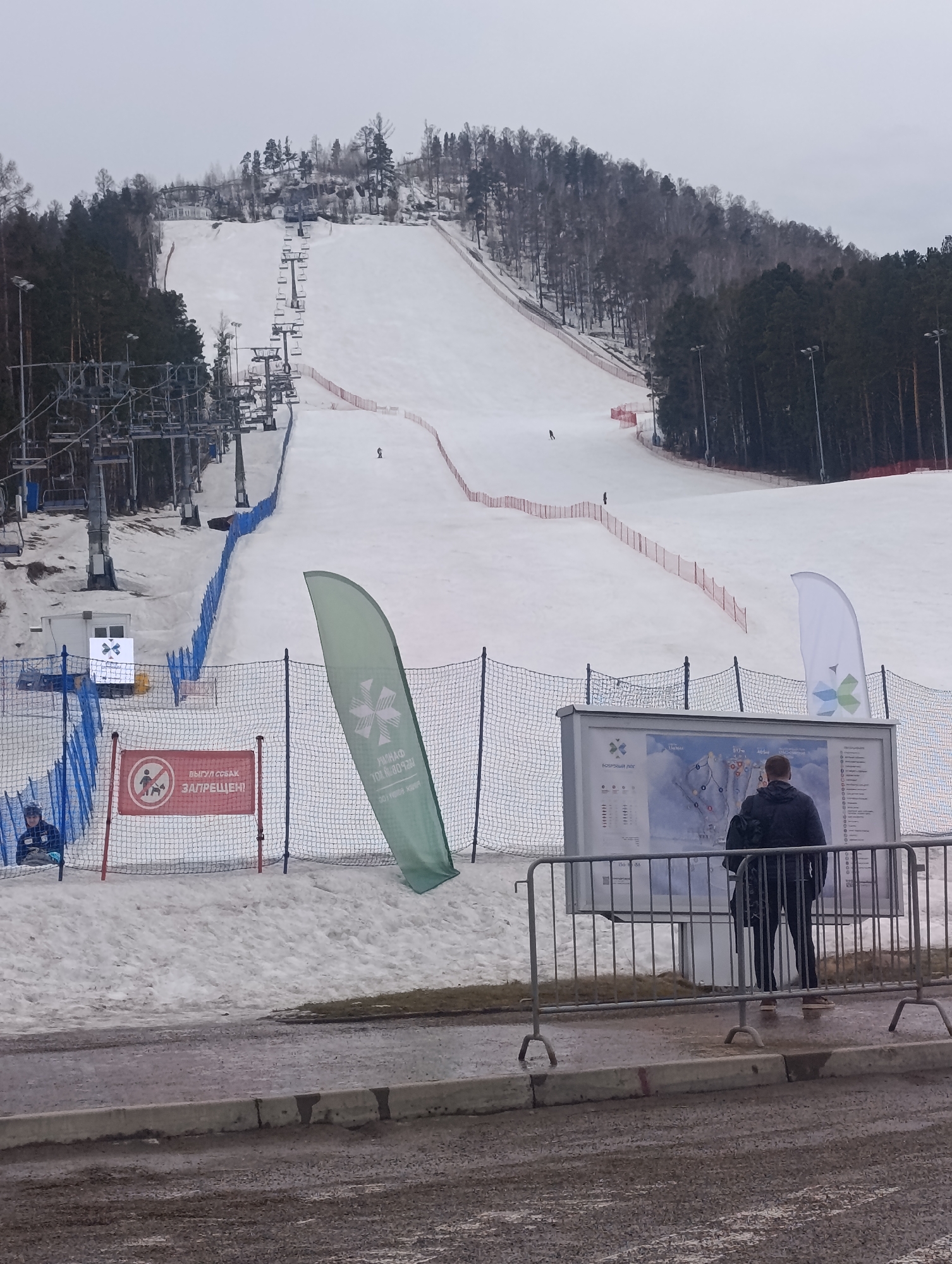 The image depicts a ski slope at the Bobrovy Log Funpark in Krasnoyarsk, Russia. A wide, snow-covered slope dominates the center of the frame, with ski lifts visible on the left. The slope is bordered by red and blue safety netting. Trees line both sides of the slope.
In the foreground, a person is standing in front of a large map, seemingly studying it. A metal fence separates the viewer from the ski slope area. Next to the map, there is a green flag with a logo on it. To the left, a red sign with Russian text and a logo is attached to the net. A person in a blue jacket is sitting behind the netting near the red sign. The ground in the immediate foreground is muddy. The sky is overcast.