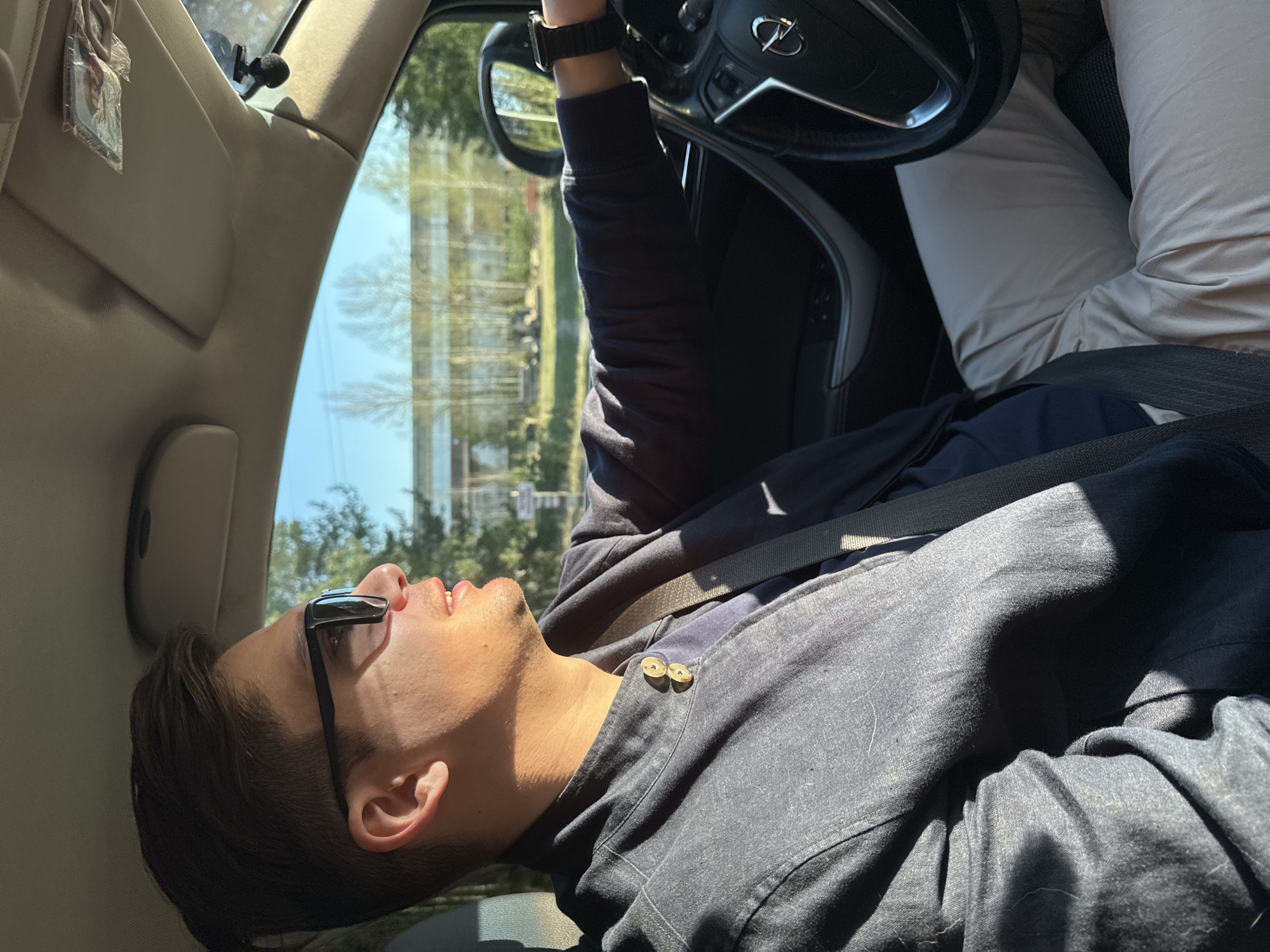 A man is reclining in the driver's seat of a car, appearing to be asleep. He is wearing sunglasses and a dark-colored button-down shirt and is fastened with a seatbelt. His hand rests on the steering wheel, which bears an Opel logo. Another person is present in the passenger seat, visible only by their clothed leg. Outside the car window, a building and trees are visible in the background. The location is Noviy, Russia.
