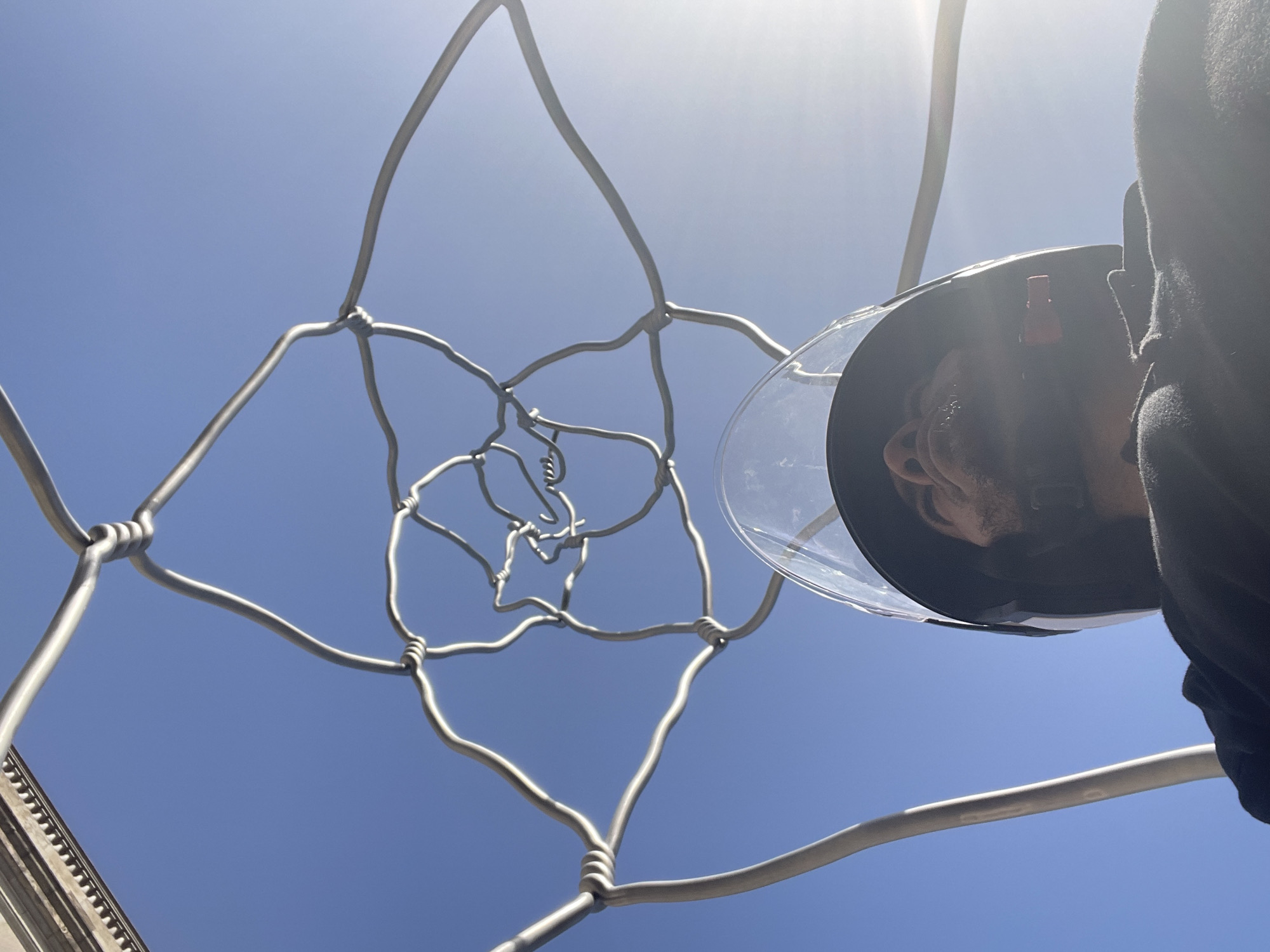 The image shows a close-up, upside-down view of a person wearing a black helmet with a clear visor, looking upwards. The person's face is partially visible through the visor. The helmet is attached to a black garment, possibly a jacket or hoodie.
Dominating the foreground is a metallic, abstract sculpture, constructed from intertwined wires. The sculpture has a complex, spiraling pattern that resembles a flower or a fractal. The wires are uniformly silver and appear to be twisted and knotted at various points to create the structure.
The background is a clear, bright blue sky, suggesting an outdoor environment. The sun is bright, casting highlights on the sculpture and the visor of the helmet. In the lower right corner of the image, a portion of an ornate building facade with architectural details, including molding and a textured surface, is visible.
The photograph appears to be taken from a low angle, looking up towards the sky. The perspective and the inclusion of the building facade suggest the location could be in a city with notable architecture. Given the style of the building, it is plausible that this scene is located in Barcelona, Spain, a city renowned for its distinctive architectural heritage. However, without further context or specific landmarks, this is an educated inference.