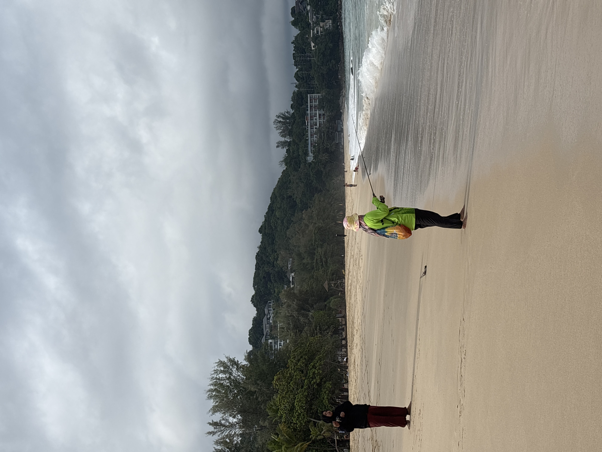The image depicts a cloudy day at a beach in Choeng Thale, Thailand. A woman in a bright green jacket, black pants, and a patterned head covering is fishing in the shallow water near the shoreline. The waves are breaking gently on the sand. Behind her, a hillside with buildings and lush green trees rises. In the foreground, another person wearing a dark hooded sweatshirt and dark red pants is standing on the beach, facing away from the water.