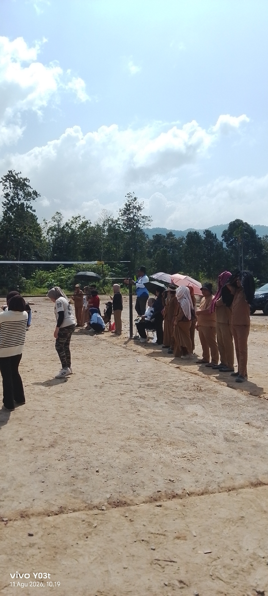 A group of people are gathered outdoors on a dirt field, likely in Tirawuta, Indonesia, given the timestamp. A volleyball net is visible in the background, suggesting the location might be a sports field or park. The weather appears to be sunny with some clouds.
In the foreground, on the left, a person wearing a striped long-sleeved shirt and black pants stands facing away from the camera. To their right and slightly behind, another person in a white t-shirt with "99" printed on it and camouflage pants is looking down. Several other individuals are seen standing and sitting in a line further back. Many are wearing brown uniforms, and some are holding umbrellas, indicating it might be a school event or a similar organized gathering. A person in a blue shirt stands near a pole, possibly overseeing the activity. Trees and a forested hillside form the backdrop. A black SUV is parked on the far right edge of the image. The ground is a light brown, sandy soil with scattered small stones.