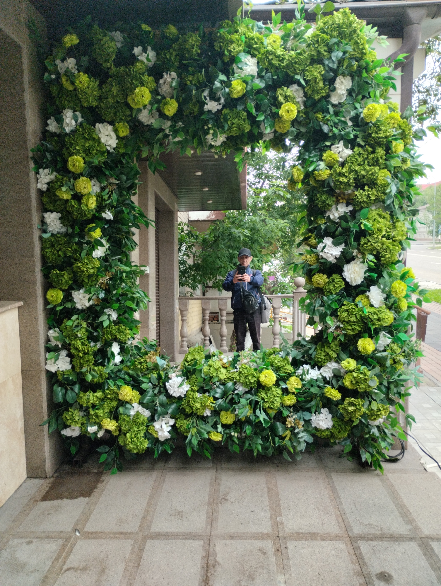 A man in a dark blue jacket and black cap stands in the center of a large, rectangular floral archway. He is holding a smartphone and appears to be taking a photo. The archway is decorated with an abundance of green foliage and clusters of white and light green artificial flowers. The man is positioned on a tiled patio, with a building entrance visible behind him and a railing to his right. The scene suggests an outdoor event or decorative installation in a city.