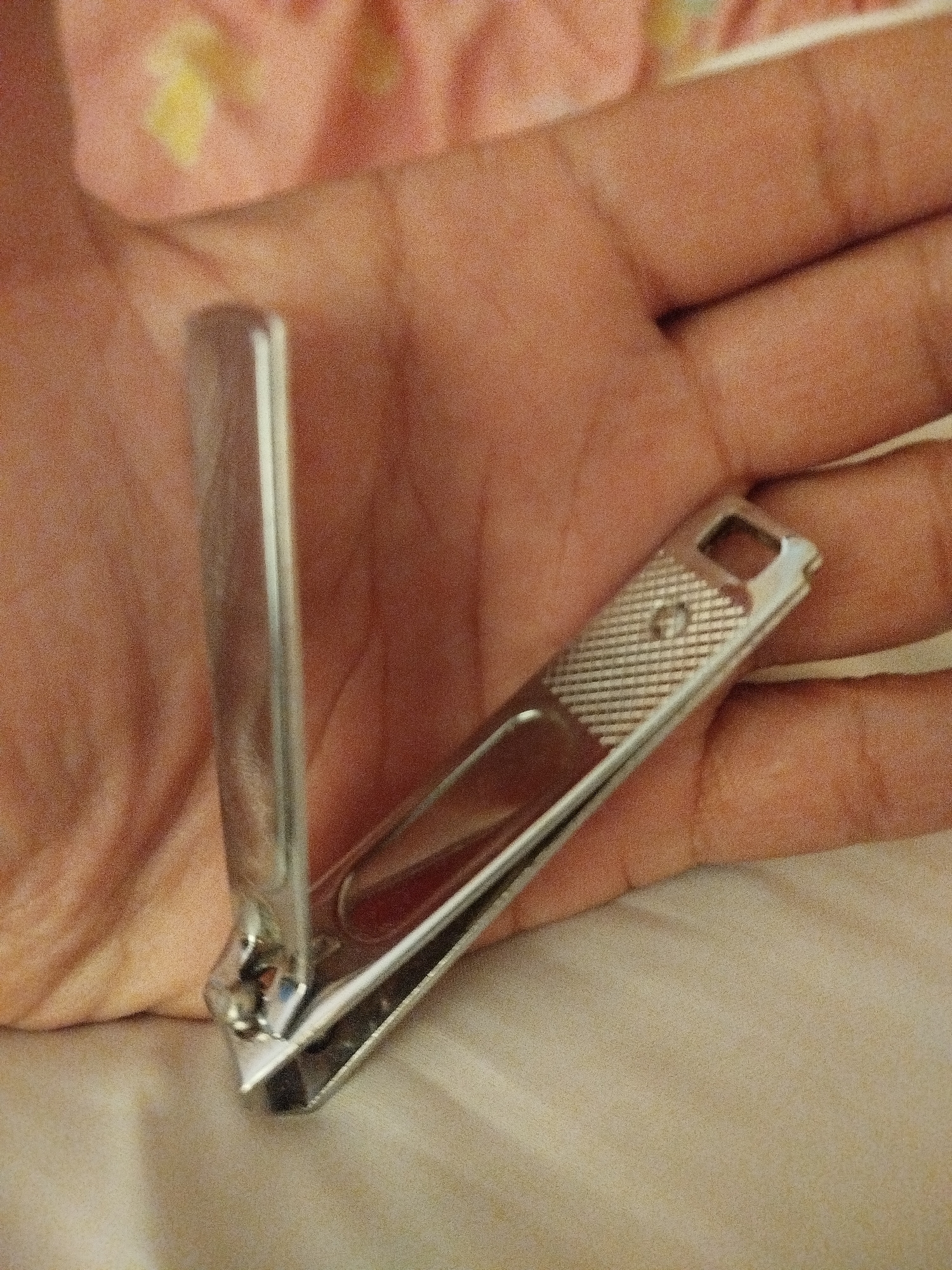 A pair of stainless steel nail clippers is held open in a person's hand. The clippers are positioned diagonally across the palm, with the lever arm extended upwards. The hand is resting on a cream-colored fabric, possibly a blanket or bedsheet, which is slightly rumpled. The background is blurred and shows a pink and yellow patterned fabric. There is no identifiable location in Lassan, Pakistan.