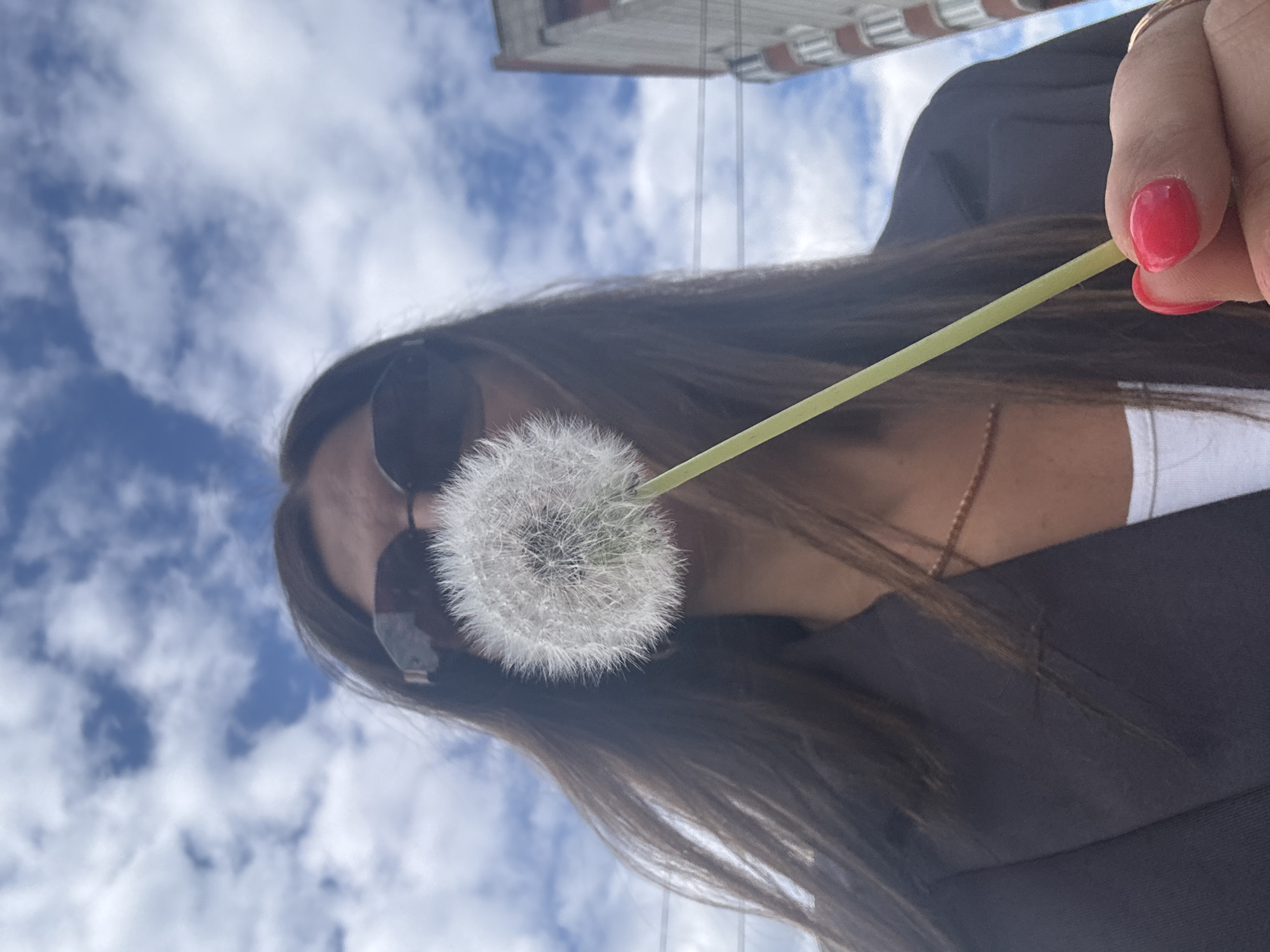 An upward-angled shot features a person, likely female, holding a fluffy white dandelion seed head against a bright blue and cloudy sky in Chaikovsky, Russia. The person, with long dark hair, wears dark sunglasses that obscure their eyes, a dark outer garment, and a white top underneath, revealing a delicate necklace. Their hand, with bright red nail polish, grasps the slender green stem of the dandelion, positioning it near their face. In the upper left background, part of a building with visible windows and several power lines are subtly visible against the expansive sky. The image captures a daytime scene with soft, scattered clouds. No text is visible in the image.