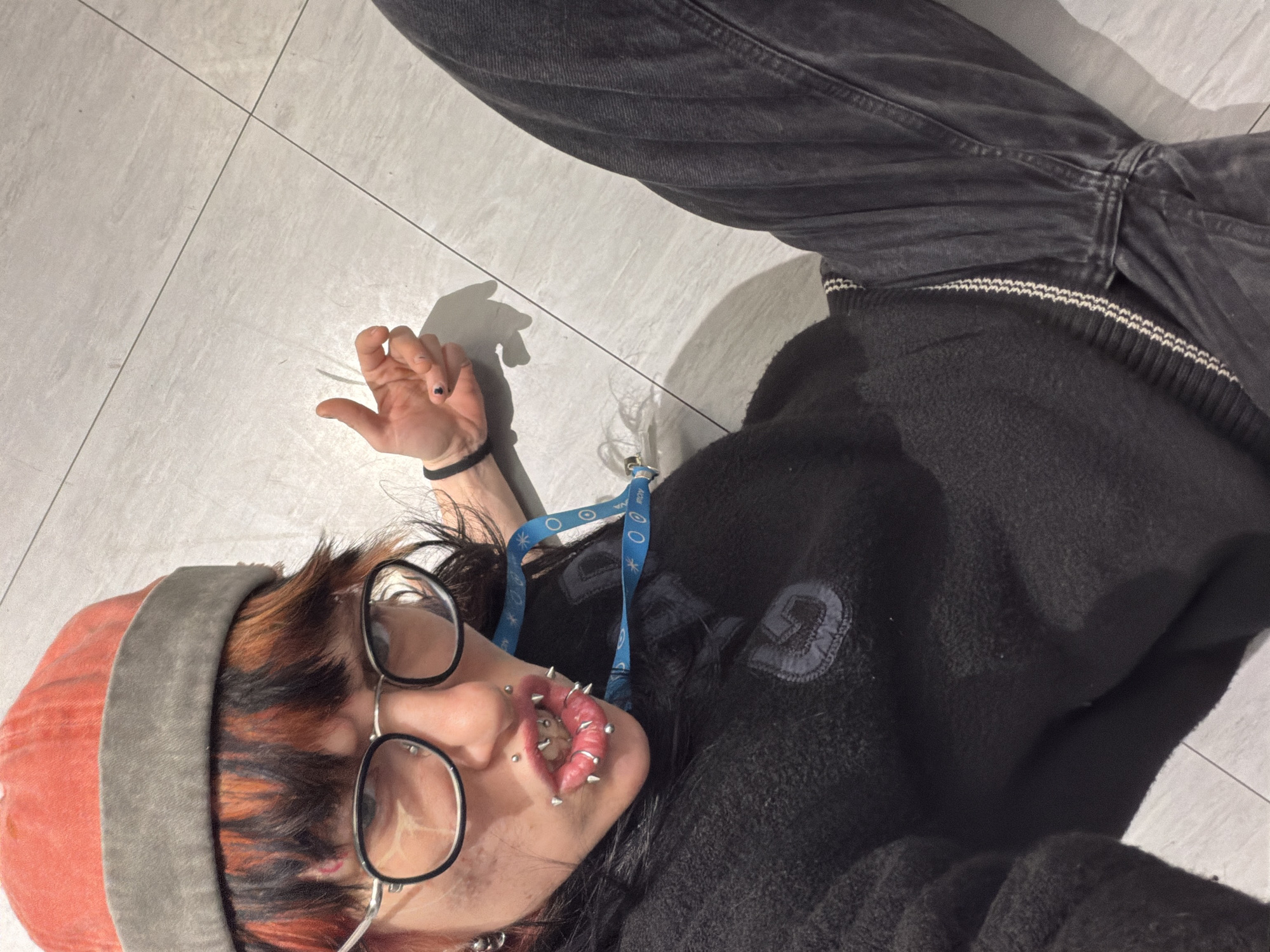 An overhead photograph captures a person lying on a light grey tiled floor, looking directly up at the camera. The individual is dressed in a black fleece jacket with a grey patch featuring an abstract 'G' or similar logo on the left chest, and dark grey jeans. They wear a reddish-orange beanie with a grey band and round-rimmed glasses. Their hair is dark with distinct reddish-orange streaks.
Notable facial features include multiple piercings: several spike-studded rings on their lips and additional nose piercings. A blue lanyard with white text and circular symbols hangs around their neck; visible text segments include "ACADEM" and "Sofia". Their left hand, showing dark nail polish on one finger and a black wristband, rests near their head. The person's expression is neutral, partially obscured by their glasses and piercings. The setting is indoors, with even, artificial lighting. The tiled floor suggests a public or institutional building in Sofia, Bulgaria, aligning with the visible text on the lanyard. The pose appears deliberate, possibly for the photograph.