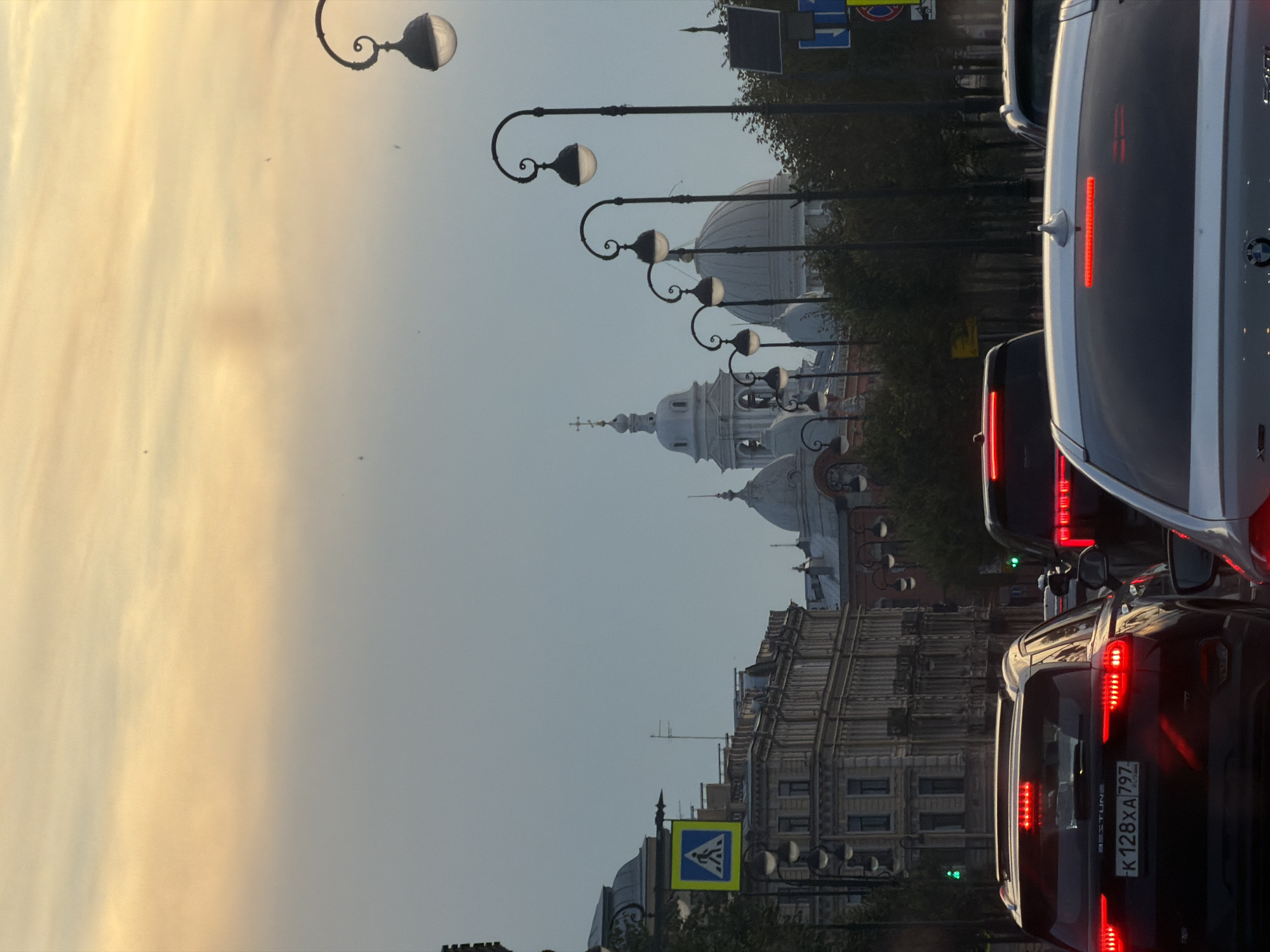 The image shows a street scene in Saint Petersburg, Russia, likely during twilight. A line of cars is stopped in traffic on a wide street. The sky is a gradient of pale yellow and grey, with a few faint clouds. On the left side of the frame, an ornate building with classical architecture and domes is visible. Streetlights with distinctive curved posts and glowing globes line the street, stretching into the distance. A pedestrian crossing sign is also present on the left. In the foreground, the taillights of several cars are illuminated. One car in the foreground has a license plate that reads "K128XA 797 RUSSIA". The overall impression is one of a busy city street at the end of the day.