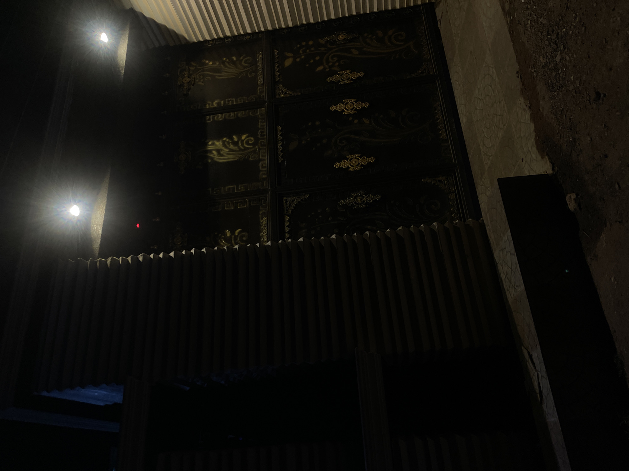 The image depicts an outdoor scene at night, possibly in Abuja, Nigeria. A large, dark gate with ornate gold-colored designs dominates the upper portion of the image. Above the gate, there is a corrugated metal awning or roof. Two bright lights are visible on the left side of the frame, casting harsh shadows. Below the gate, a slatted wooden structure partially obscures the scene, suggesting a fence or enclosure. To the right of the gate, there is a rough, unpaved ground surface. The overall scene is dark and visibility is limited.
