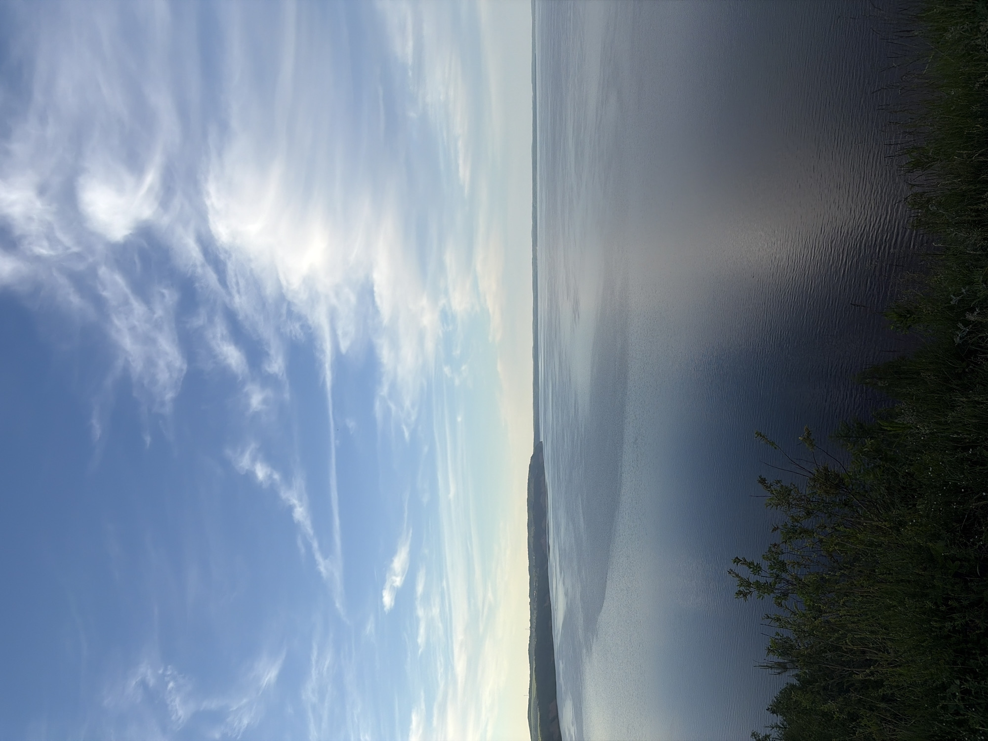 The image captures a vast and serene natural landscape, dominated by an expansive sky and a large body of water. The horizon line, formed by a low, distant landmass, stretches horizontally across the middle of the frame, separating the sky from the water.
The upper half of the image is filled with a bright, light blue sky adorned with delicate, wispy white clouds, possibly cirrus or cirrocumulus, which are stretched across the expanse, some appearing brighter due to sunlight. A distinct bright area in the sky suggests the position of the sun, casting a soft illumination over the scene.
Below the horizon, a wide body of water, likely a large lake or river, extends into the distance. The water's surface exhibits subtle ripples, reflecting the sky's light and colors. The water appears darker in the foreground, transitioning to lighter tones towards the horizon. The distant landmass forming the horizon appears low and indistinct, possibly covered with greenery.
In the immediate foreground on the right side, dark green foliage, consisting of bushes or trees, frames a portion of the water, indicating the image was captured from an elevated position overlooking the water body.
The scene conveys a sense of tranquility and open space. The time of day appears to be during daylight hours, perhaps late morning or afternoon, given the clear sky and diffused sunlight. The weather is calm, with light clouds and still water. No people or specific activities are visible. No text is present in the image. The setting is consistent with a natural environment near a significant water feature in Костоваты, Russia.