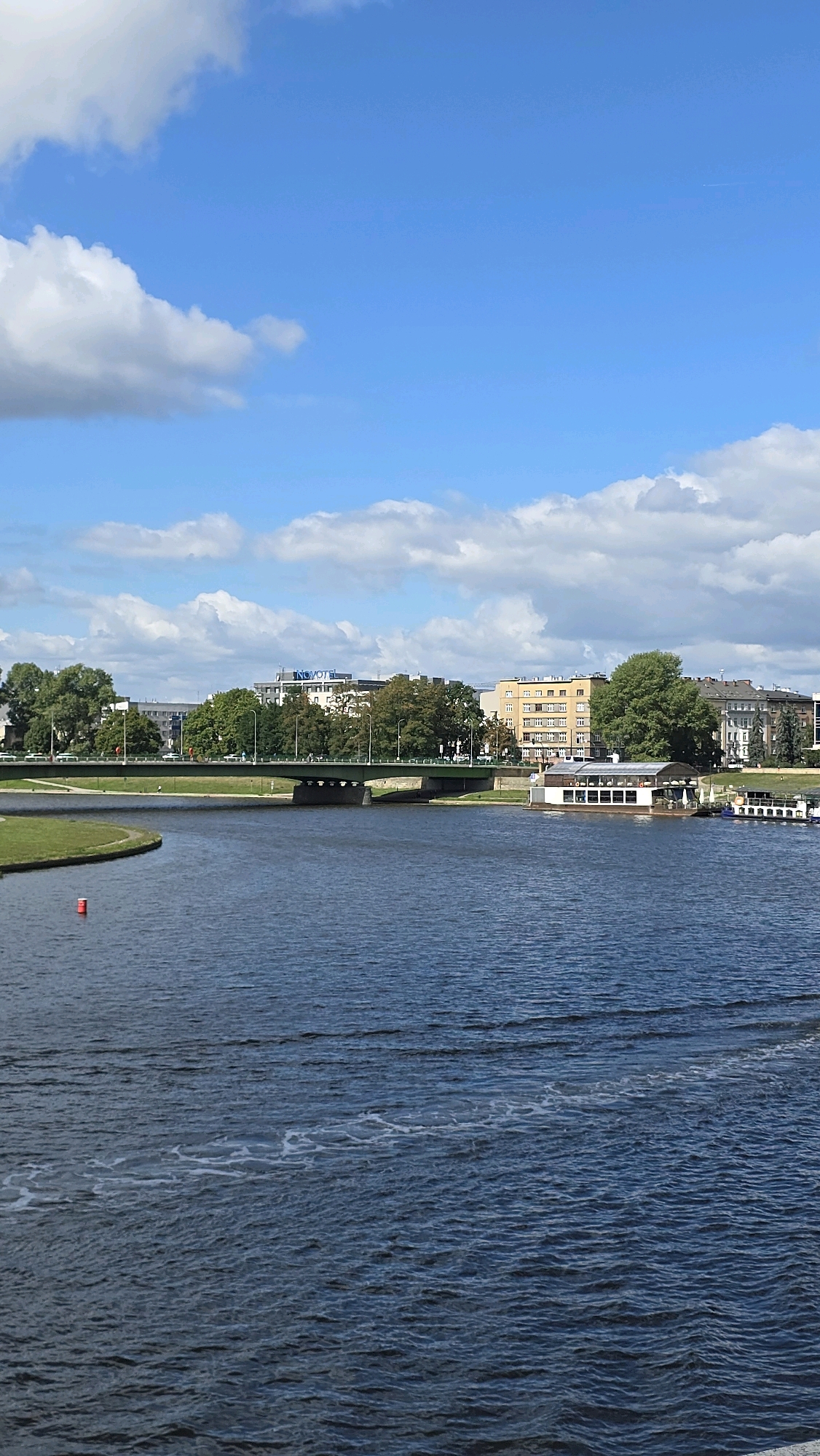 The image shows a panoramic view of the Vistula River in Krakow, Poland. The river occupies the foreground and mid-ground of the image, with gentle ripples disturbing its surface. On the left bank of the river, a small, circular red buoy is visible in the water.
A green bridge spans the river in the mid-ground, with several cars crossing it. Beyond the bridge, a line of trees separates the riverfront from the city skyline. Several buildings are visible, including a modern hotel with a prominent "NOVOTEL" sign. To the right of the bridge, a white building with large windows, possibly a restaurant or cafe situated on the river, is visible. A boat is docked next to this building. Further in the background, more buildings and trees form the urban landscape.
The sky above is a vibrant blue, dotted with fluffy white clouds, indicating fair weather. The scene is bathed in sunlight, casting soft shadows and highlighting the colors of the environment. The overall impression is one of a calm, sunny day in a city by the river.