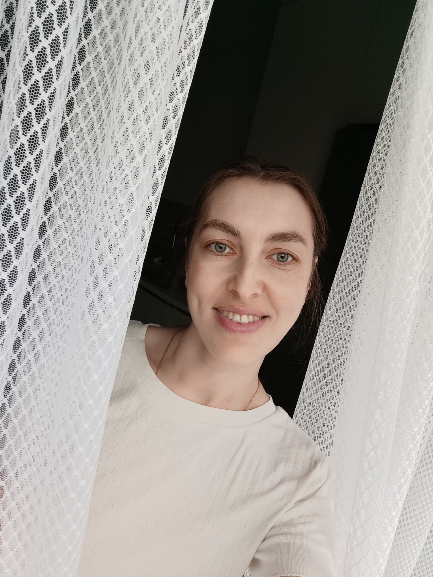 A woman is standing behind white lace curtains. She has fair skin, light brown hair pulled back, and is wearing a cream-colored t-shirt. She is smiling and looking directly at the camera. Behind the curtains is a dark space, possibly a room. The location is Chaikovsky, Russia.