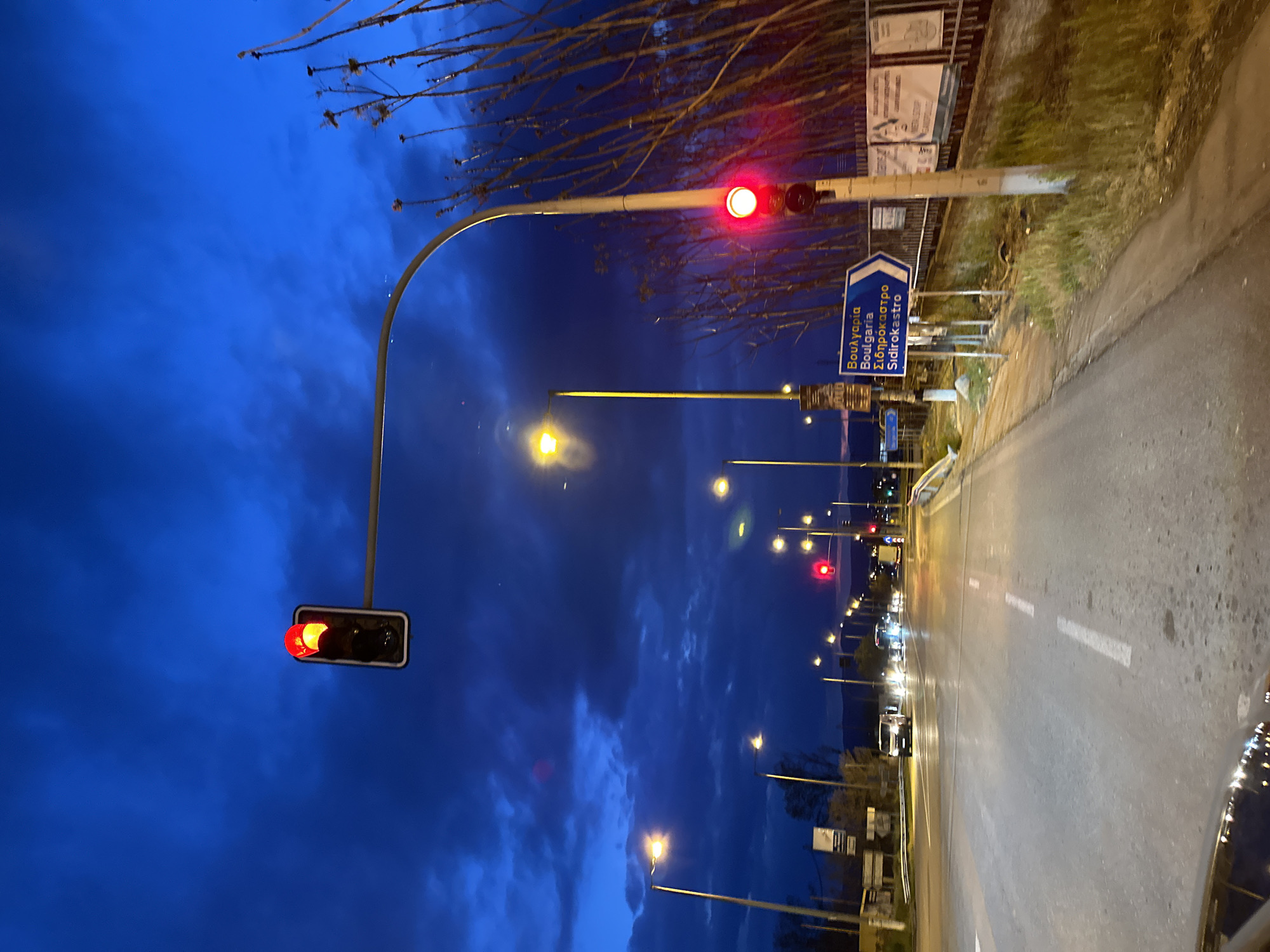 The image captures a street scene at dusk in Lefkonas, Greece. A red traffic light hangs on a curved metal support over the road. Streetlights illuminate the asphalt, which is marked with white lines. A blue road sign points towards Bulgaria and Sidirokastro. Several cars with headlights on are visible in the distance. The sky is a deep blue with dark clouds. Bare tree branches extend into the upper part of the frame. There are buildings and other signage lining the side of the street.