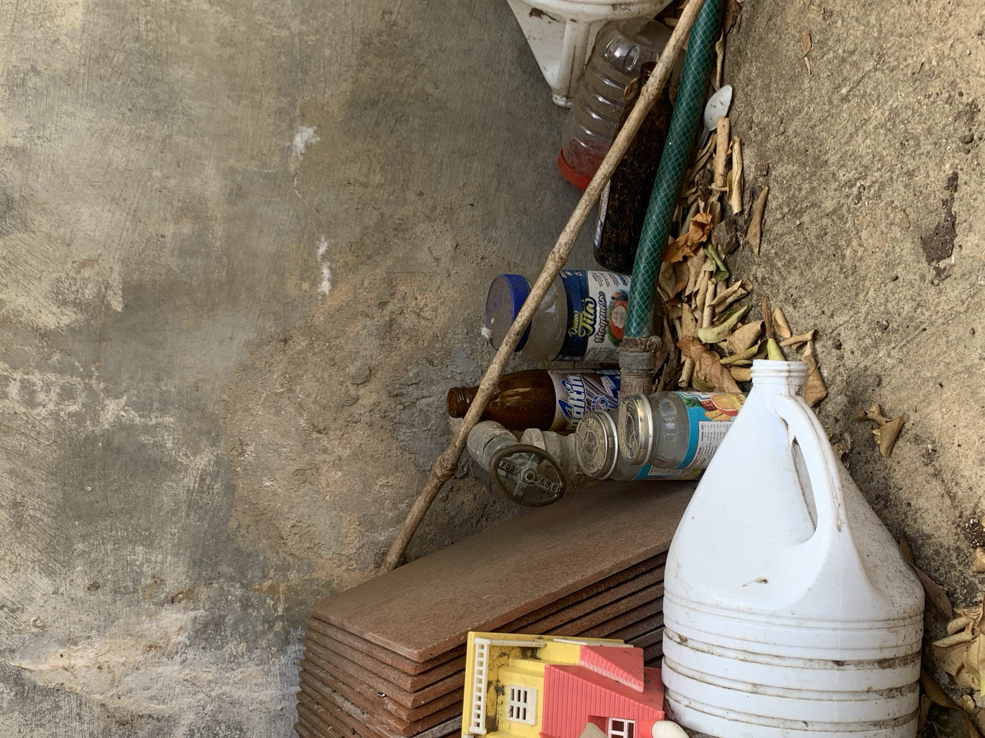 The image presents a cluttered, static scene in what appears to be an outdoor utility or storage area, likely in Santa Ana, Venezuela. The setting is dominated by concrete surfaces, including a rough ground and a textured wall.
Various objects are scattered or stored in the frame. In the top right, a white ceramic object, possibly a part of a sink or toilet, is partially visible. Below it, several plastic bottles, including a clear one with a red cap and a blue-capped bottle, are present. The blue-capped bottle clearly displays the text "Tito's" and "Margaritas". Another brown glass bottle, partially obscured, shows the brand "Maltin". A green garden hose runs vertically along the right side, partially covered by dry leaves and debris that accumulate against the concrete wall and ground. A wooden stick or rod lies diagonally across the image.
Towards the bottom center, a stack of reddish-brown rectangular objects, possibly tiles or planks, is neatly arranged. Resting near these items is a miniature building model, yellow with a red roof, featuring small white window frames and a ladder. In the bottom right corner, a large white plastic jug with a handle stands upright, appearing dirty. Near the center, a metal valve or pipe fitting is visible amidst other items. A can with the word "Premium" is also partly visible.
No people are present, and no activity is taking place. The lighting suggests it is daytime, with natural light illuminating the scene. The overall impression is one of disarray and neglect in a functional but untidy space.