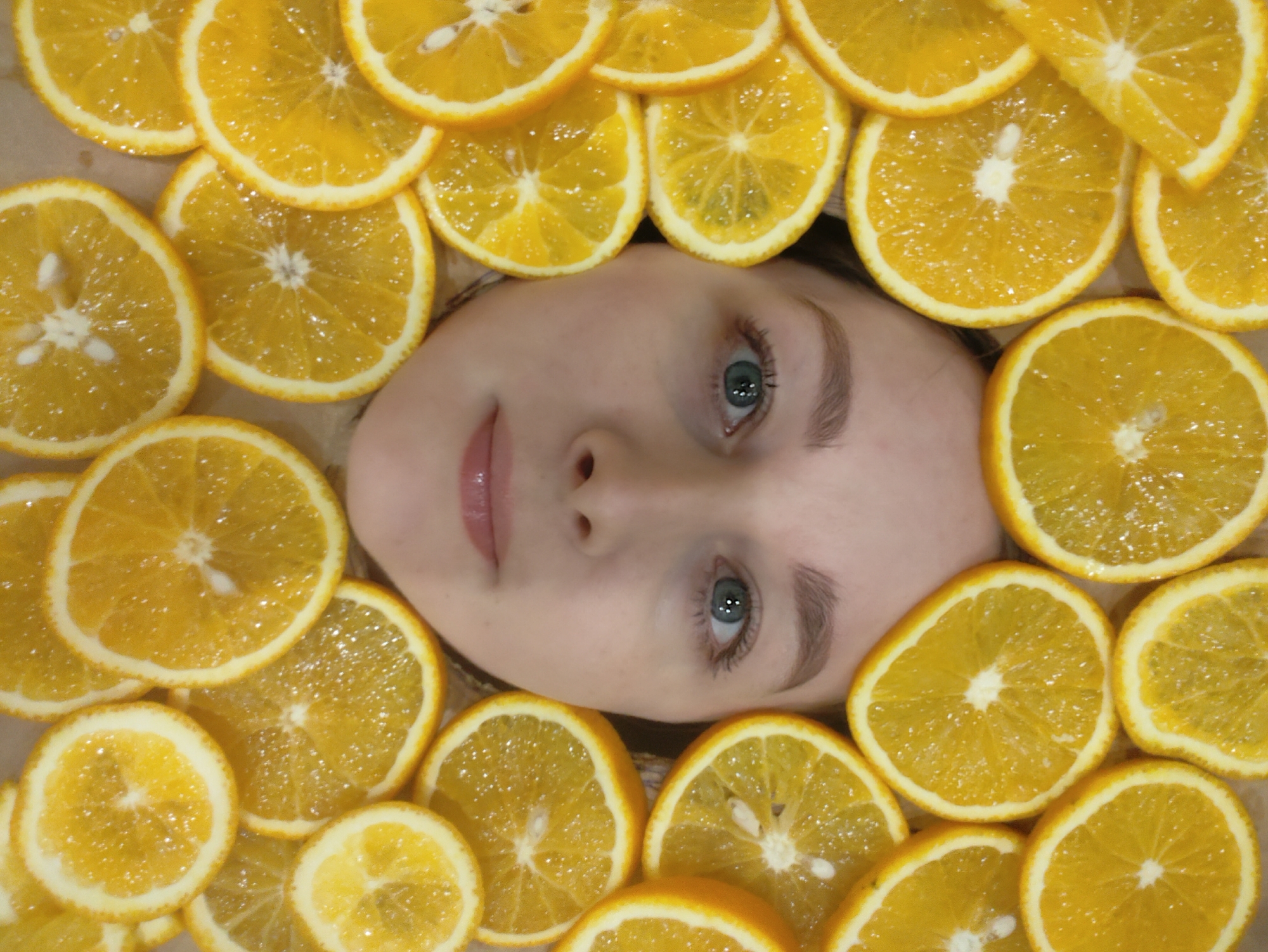 A close-up, eye-level shot shows a person's face surrounded by orange slices. The face is centered, with the slices forming a border. The person has fair skin, light eyebrows, and light blue eyes. The orange slices are mostly complete circles, with some partial slices around the edges. The slices appear fresh and juicy. The background is a neutral color, possibly light brown or beige. The location is unspecified.