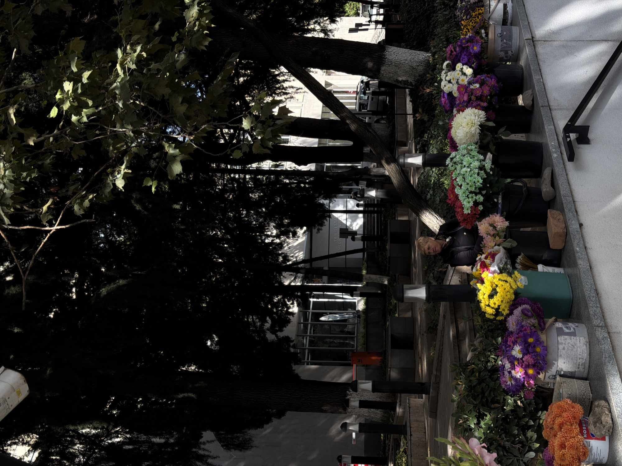 A woman is standing behind a display of flowers, seemingly for sale, in an outdoor urban setting. The flowers, in various colors and arrangements, are placed in buckets and containers on a ledge. The woman, with short blonde hair and wearing a dark jacket, appears to be observing her surroundings. Behind the flower display, trees with dark foliage provide shade. In the background, a building with large windows and some parked vehicles can be seen. The scene suggests a street-level market or flower stall in a city. The presence of building architecture and the sign in the background indicating "TBILISI STATE..." suggests the location is in Tbilisi, Georgia.
