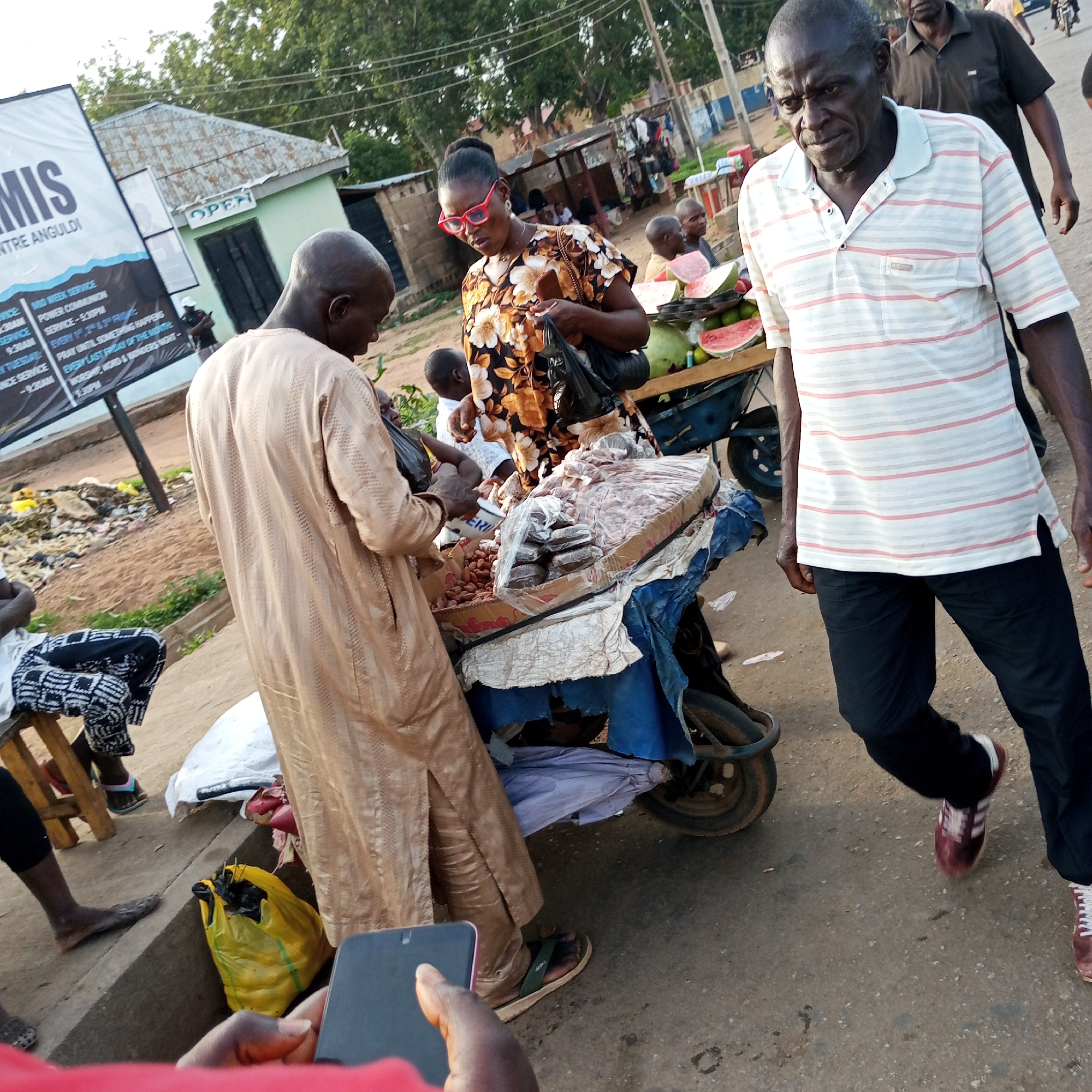 A daytime street scene in Bukuru, Nigeria, shows several people and vendors along a road. In the foreground, a man in a horizontal-striped shirt and black pants walks toward the camera, a cart partially visible behind him. Another man in a long, tan robe stands at a cart, interacting with a woman in a brown floral dress and red glasses. The cart is laden with what appear to be food items. In the background, more people, including a person selling watermelons from another cart. To the left, a sign for "MIS CENTRE ANGULDI" is visible, and a building with an "OPEN" sign. A yellow plastic bag rests on the ground. A hand holding a smartphone can be seen at the bottom of the image.