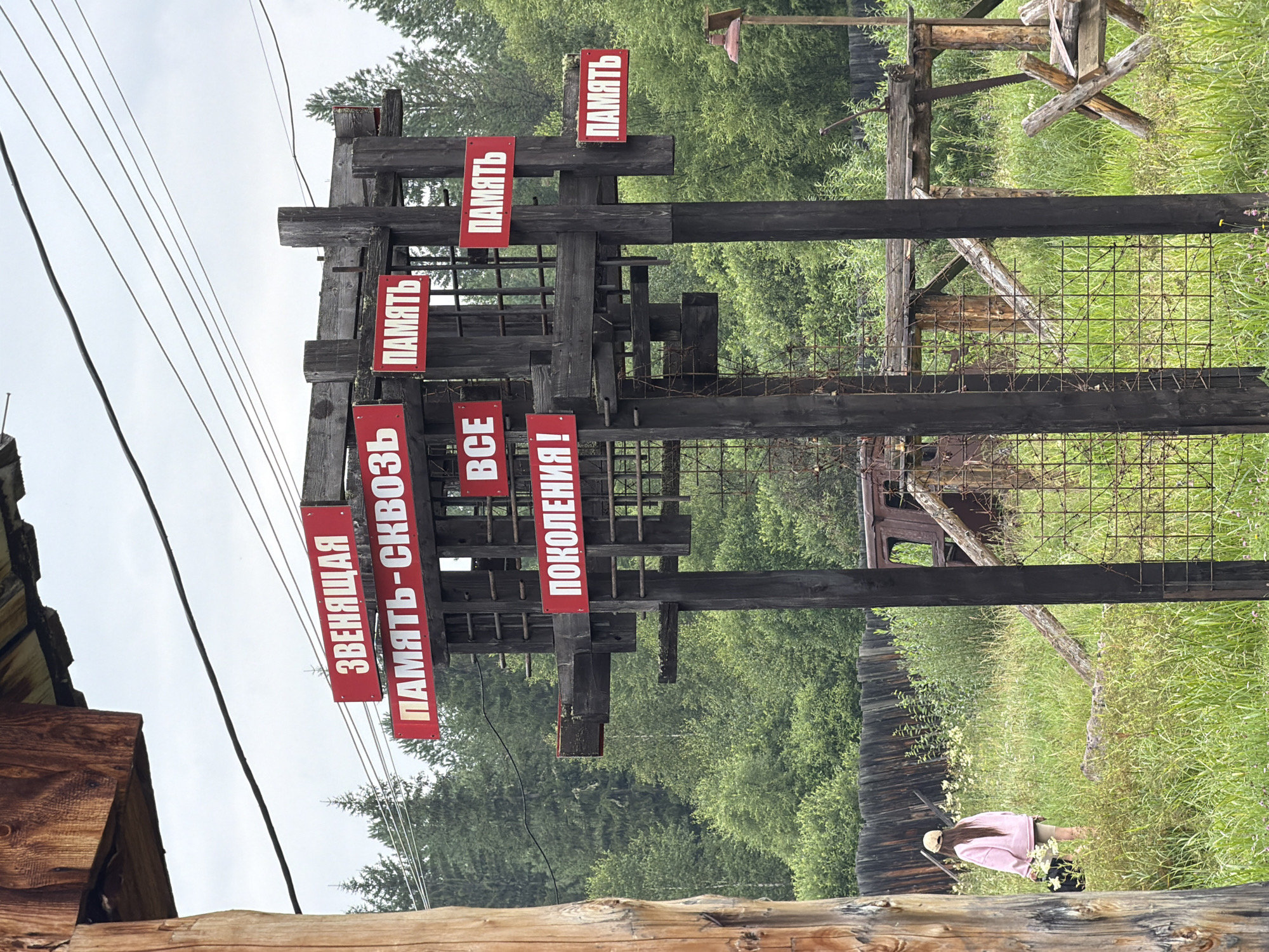 The image depicts an outdoor scene in Ust'-Kabyrza, Russia, featuring a weathered wooden structure with multiple red signs bearing white Cyrillic lettering. The signs appear to be historical or memorial in nature, with phrases like "ЗВЕНЯЩАЯ ПАМЯТЬ-СКВОЗЬ ПОКОЛЕНИЯ!" (Ringing Memory Through Generations!) and "ПАМЯТЬ" (Memory) visible. The structure is set against a backdrop of lush green trees and vegetation. In the foreground, a young person wearing a pink hoodie and shorts walks away from the camera, carrying a dark bag. The ground is covered in tall grass and wildflowers. In the background, there are remnants of what looks like an old vehicle and a rustic wooden fence. The overall atmosphere is one of historical reflection amidst a natural, rural setting.