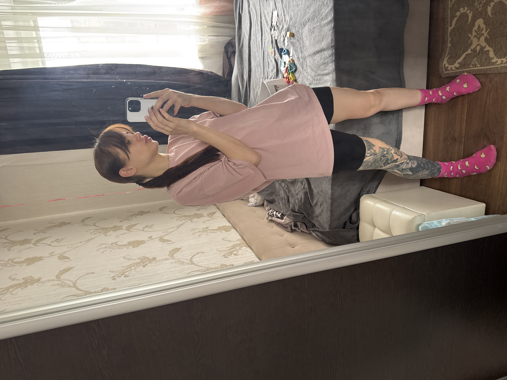The image shows a reflection in a mirror, likely within a residential space in Rostov-on-Don, Russia. A woman is captured holding a smartphone and taking a picture of herself. She is wearing a pink T-shirt and black shorts, with pink socks decorated with bananas. One leg has a visible tattoo. The background includes a bed covered with a gray sheet, various objects scattered on the bed, a white ottoman, and a portion of the room's wall with patterned wallpaper. The scene is generally indoors.