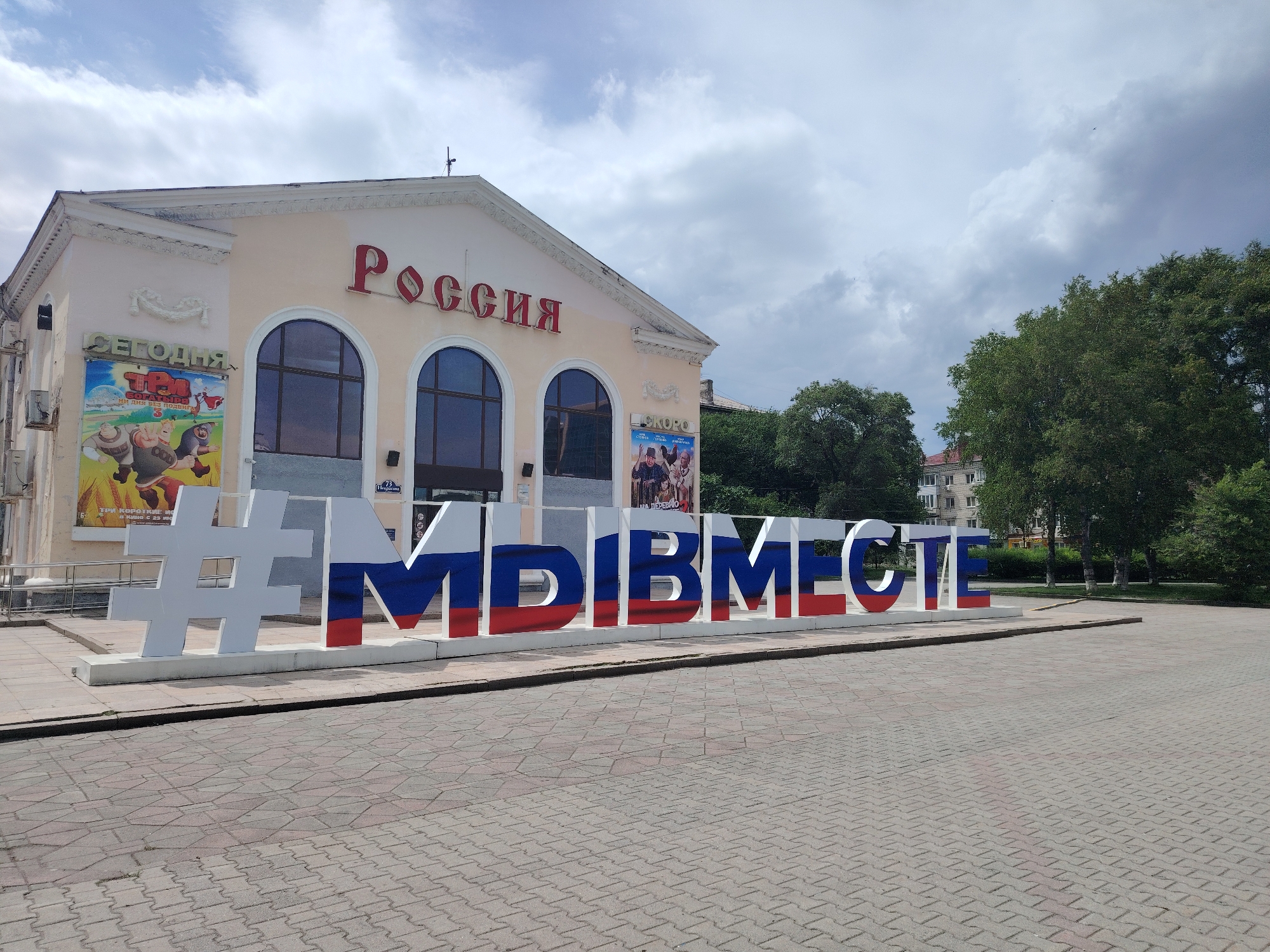 The image shows a large, three-dimensional hashtag sign in front of a building. The building has the word "РОССИЯ" (Russia) written in large red letters across its facade, and appears to be a cinema or cultural center. The hashtag sign itself is made of large, white, blocky letters, with the last four letters ("МЫВМЕСТЕ" - We are together) painted in the colors of the Russian flag: white, blue, and red. The sign is placed on a paved plaza. Behind the building, there are trees and other urban structures visible. The sky is cloudy. The location is identified as Ussurijsk, Russia, and specifically points to the "Russia" cinema.