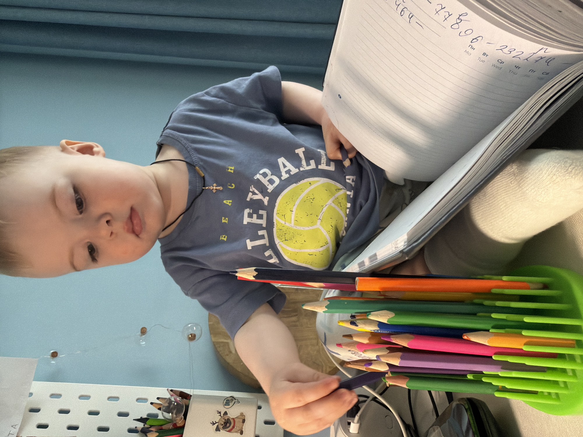 A child, approximately two years old, wearing a blue t-shirt with a yellow volleyball graphic and a dark necklace with a cross pendant, sits at a desk. The child is holding a dark purple colored pencil in their right hand and is surrounded by other colored pencils in a green container. A notebook is open on the desk. The notebook page contains handwriting in Cyrillic. The background is a light blue wall with decorative clear glass balls attached. A white shelf is visible with various items, including other pencils and a decorative item with a cartoon pug wearing a reindeer costume. The location appears to be indoors, possibly a home in Votkinsk, Russia.