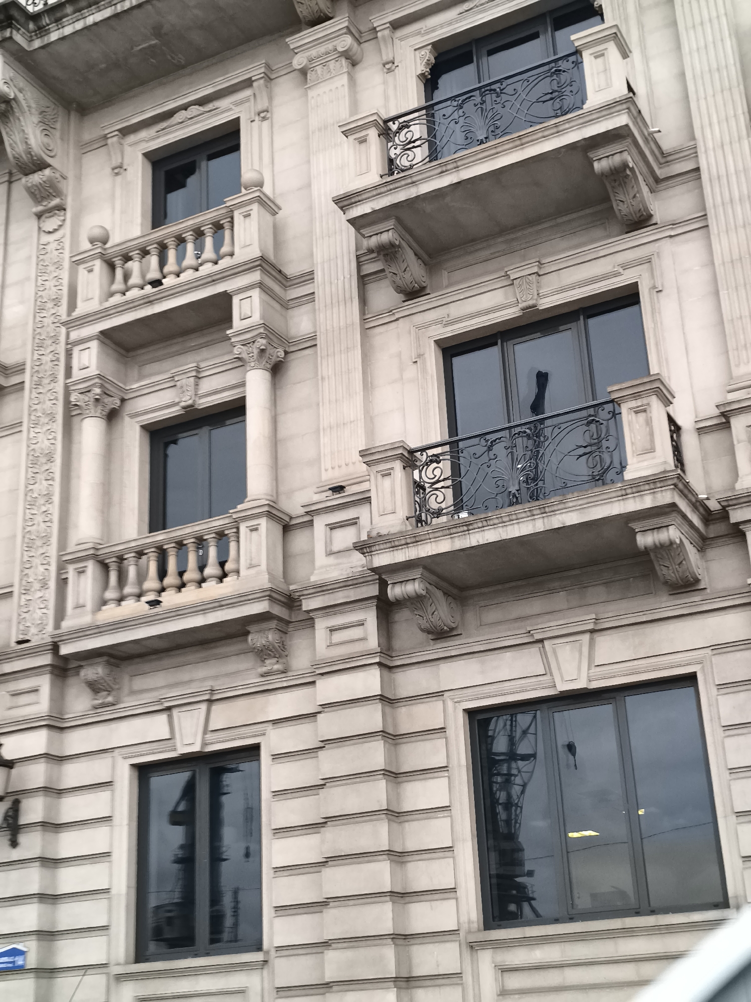 The image shows a close-up, low-angle view of a cream-colored, ornate stone building facade in Batumi, Georgia. The building features multiple levels with balconies, windows, and decorative architectural elements such as columns, ornate carvings, and moldings.
The balconies have stone balustrades with spherical finials and decorative wrought-iron railings with intricate scrollwork. The windows are large and dark-tinted, reflecting the sky and surrounding structures, including what appears to be a construction crane. Some windows have reflective surfaces that show a silhouette of a dark object hanging from the ceiling, possibly a light fixture or decorative item.
The stone facade is characterized by horizontal banding and vertical pilasters. The overall impression is one of classical or neoclassical architectural style. There are no people visible in the image. The sky appears overcast.