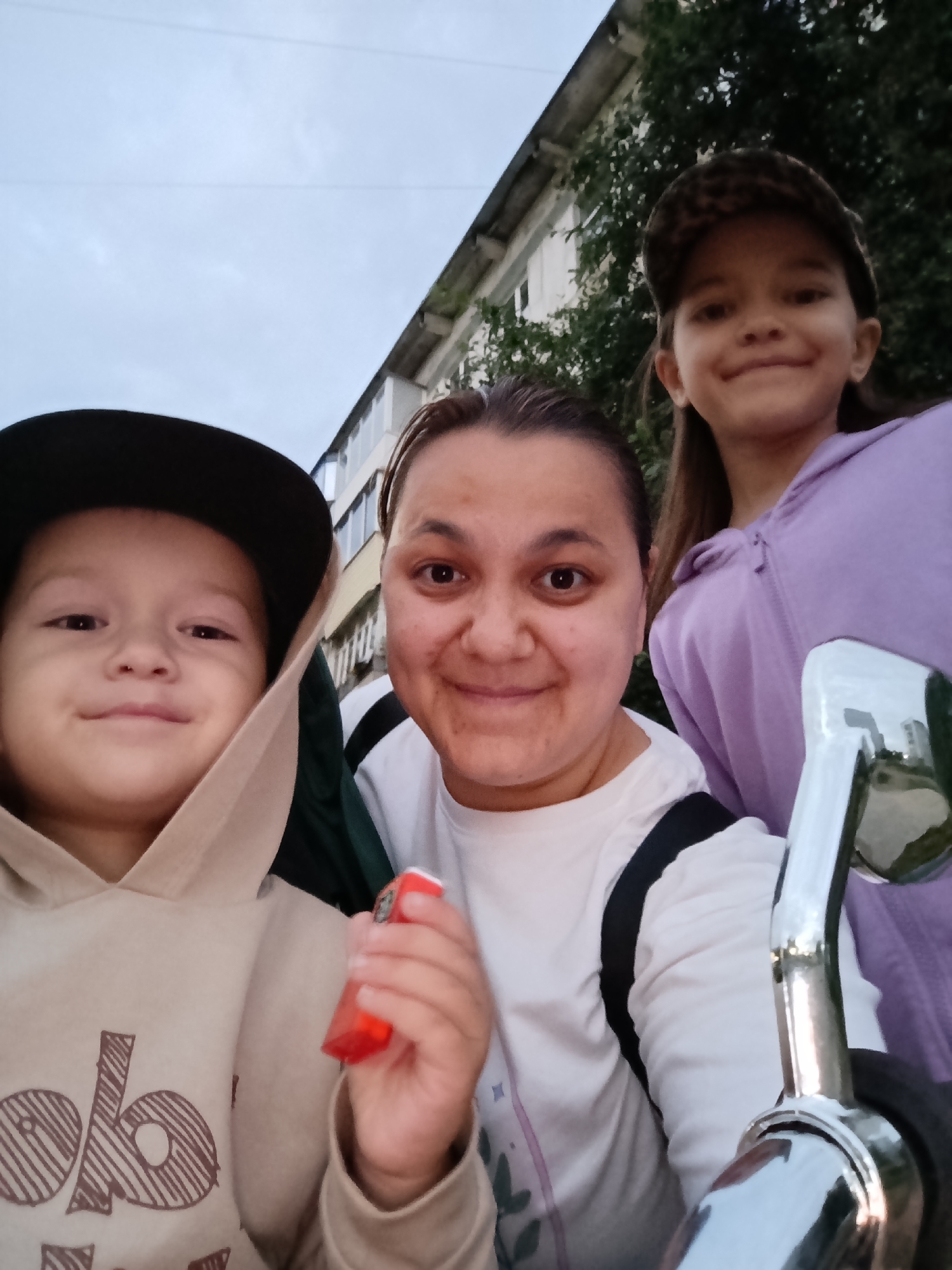 An outdoor selfie shows three individuals, two children and one adult, with a building and trees in the background. A young boy in a beige hooded sweatshirt and a black baseball cap is on the left. To his right, a woman with dark hair, wearing a white shirt, is in the center. On the far right, a young girl with long dark hair, wearing a leopard-print baseball cap and a purple hooded sweatshirt, is visible. The woman is holding a small red object. The background features a multi-story building and foliage. The overall scene suggests a casual outdoor setting in Chaikovsky, Russia, possibly on a street or in a park.
