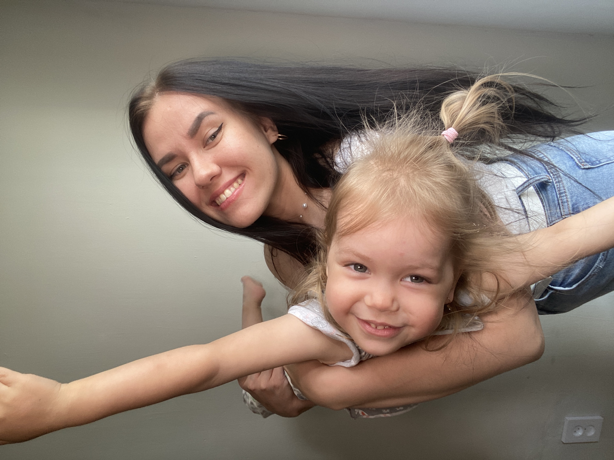 The media file shows a young woman with long, dark hair and a young girl with blonde hair. The woman is holding the girl, who is facing the camera with a wide smile. The girl has her arms around the woman's neck. The woman is also smiling. Both subjects are wearing casual clothing; the woman appears to be wearing a white top, and the girl is wearing a white top with a pattern. The background is a plain, light-colored wall. The lighting suggests it is daytime, possibly indoors. The overall mood is happy and playful. There is a visible electrical outlet on the wall in the lower right corner of the image. The location context is Chaikovsky, Russia.