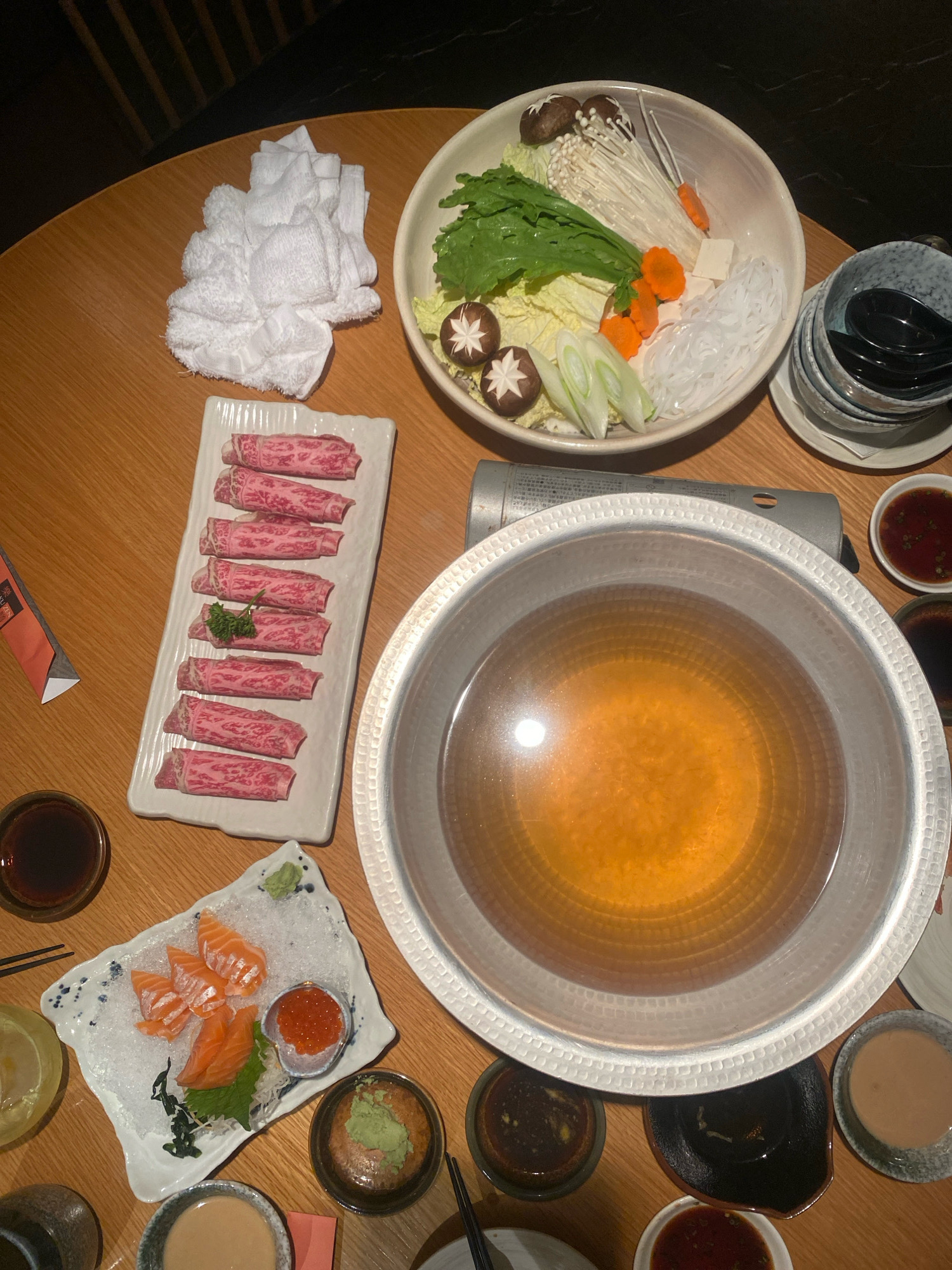 This is a top-down, eye-level photograph of a shabu-shabu meal set on a wooden table. The meal includes thinly sliced raw beef, salmon sashimi with roe, various vegetables such as napa cabbage, mushrooms, enoki mushrooms, and scallions, clear noodles, and a pot of broth heated by a portable stove. There are also several small dishes of dipping sauces. The table is set in what appears to be a restaurant setting, with a blurred background that suggests an indoor environment. The lighting is warm and ambient. The image depicts a meal ready to be cooked and eaten. This scene is consistent with dining experiences found in various restaurants across Kuala Lumpur, Malaysia, particularly those specializing in Japanese cuisine.