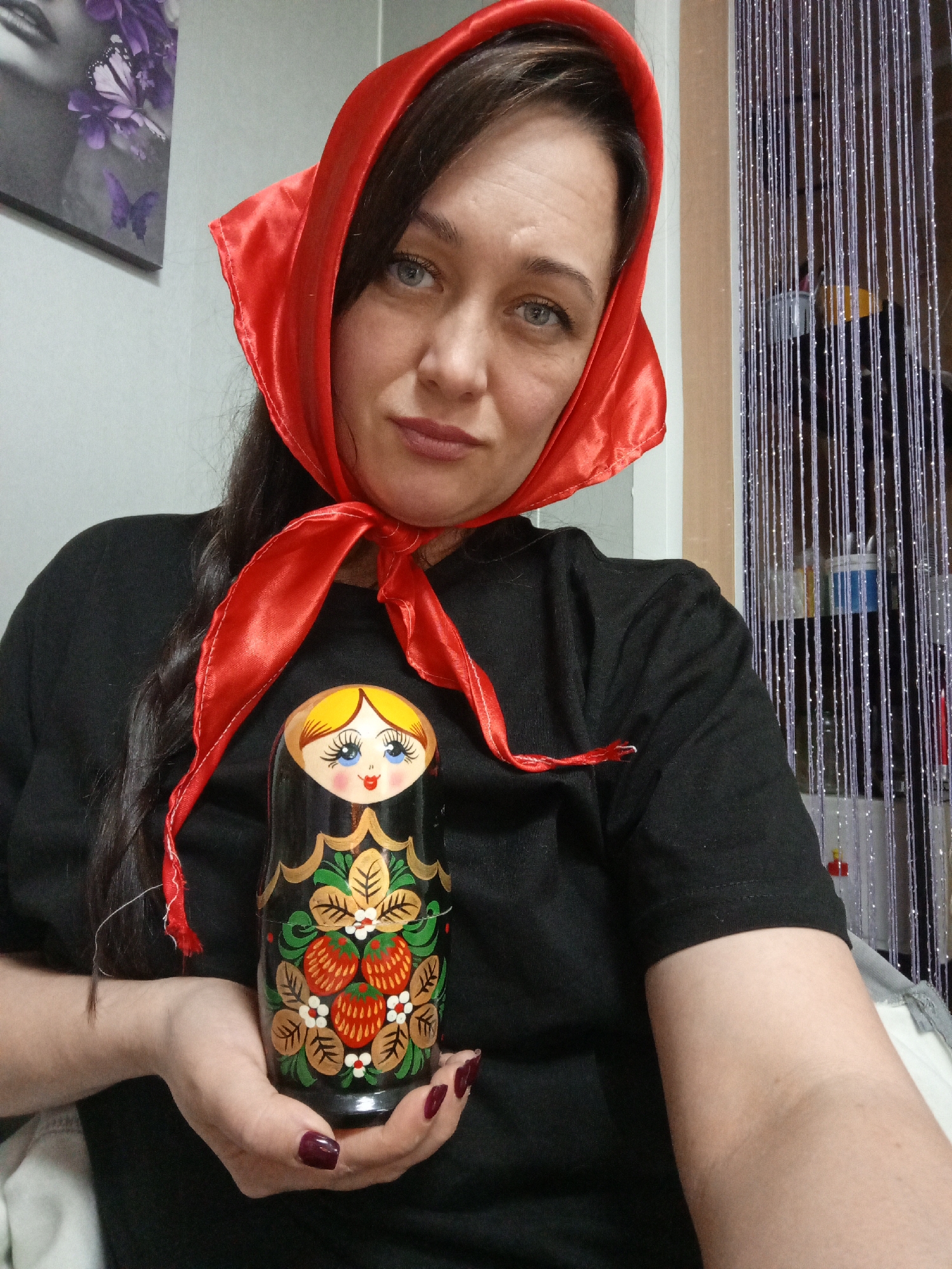 A woman with long dark hair and striking blue eyes is depicted indoors, wearing a bright red satin headscarf tied beneath her chin and a black t-shirt. She holds a traditional Russian Matryoshka nesting doll, featuring a black base adorned with a cheerful face, blonde hair, and an intricate floral and strawberry motif. Her fingernails are painted a dark red. The woman looks directly at the camera with a calm, subtle expression.
The background to her left features a light grey wall with a partial view of a black and white artwork depicting a woman's face and purple butterflies. To her right, a shimmering beaded curtain, likely silver or light purple, hangs vertically, partially obscuring shelves or storage units behind it. The indoor lighting suggests the photograph was taken without strong natural light. The presence of the Matryoshka doll and the headscarf, often associated with Russian culture, aligns with the given context of Chaikovsky, Russia, suggesting a personal setting such as a home. There is no visible text in the image.