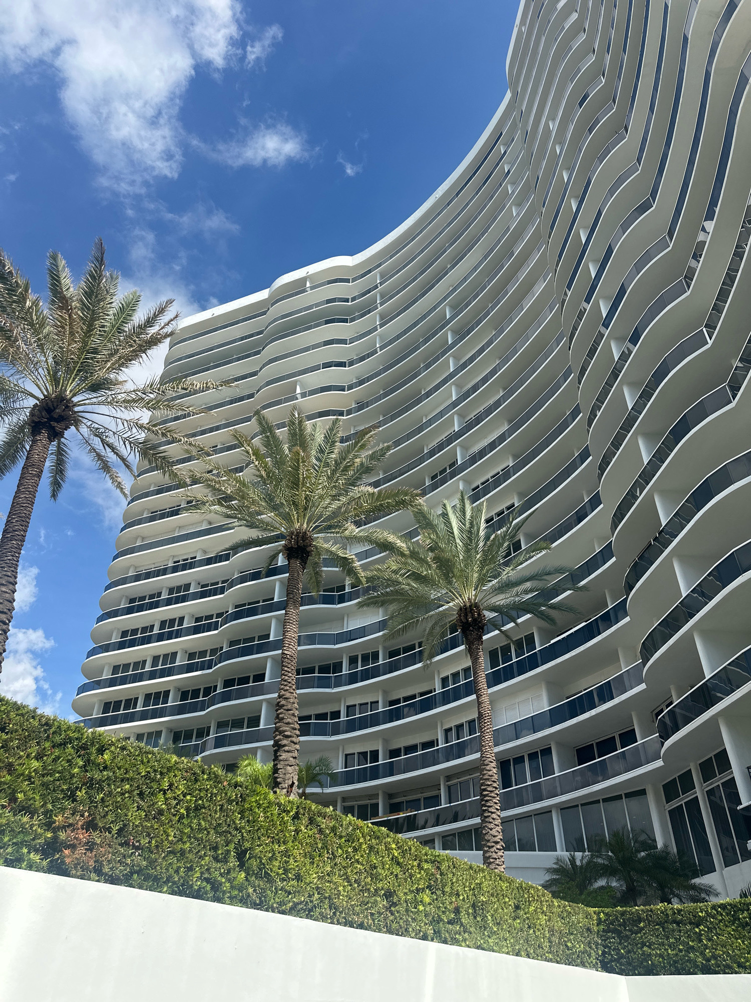 A modern, white, curved apartment building with many balconies stands tall against a bright blue sky with scattered white clouds. Three palm trees are in the foreground, with lush green bushes and a white wall below them. The building's balconies are defined by undulating white railings and dark glass panels, creating a wave-like pattern. The architecture suggests a location in a warm, sunny climate, likely near a coast, fitting for Surfside, Florida. The image is taken from a low angle, emphasizing the scale and grandeur of the building. No people are visible in the scene.
