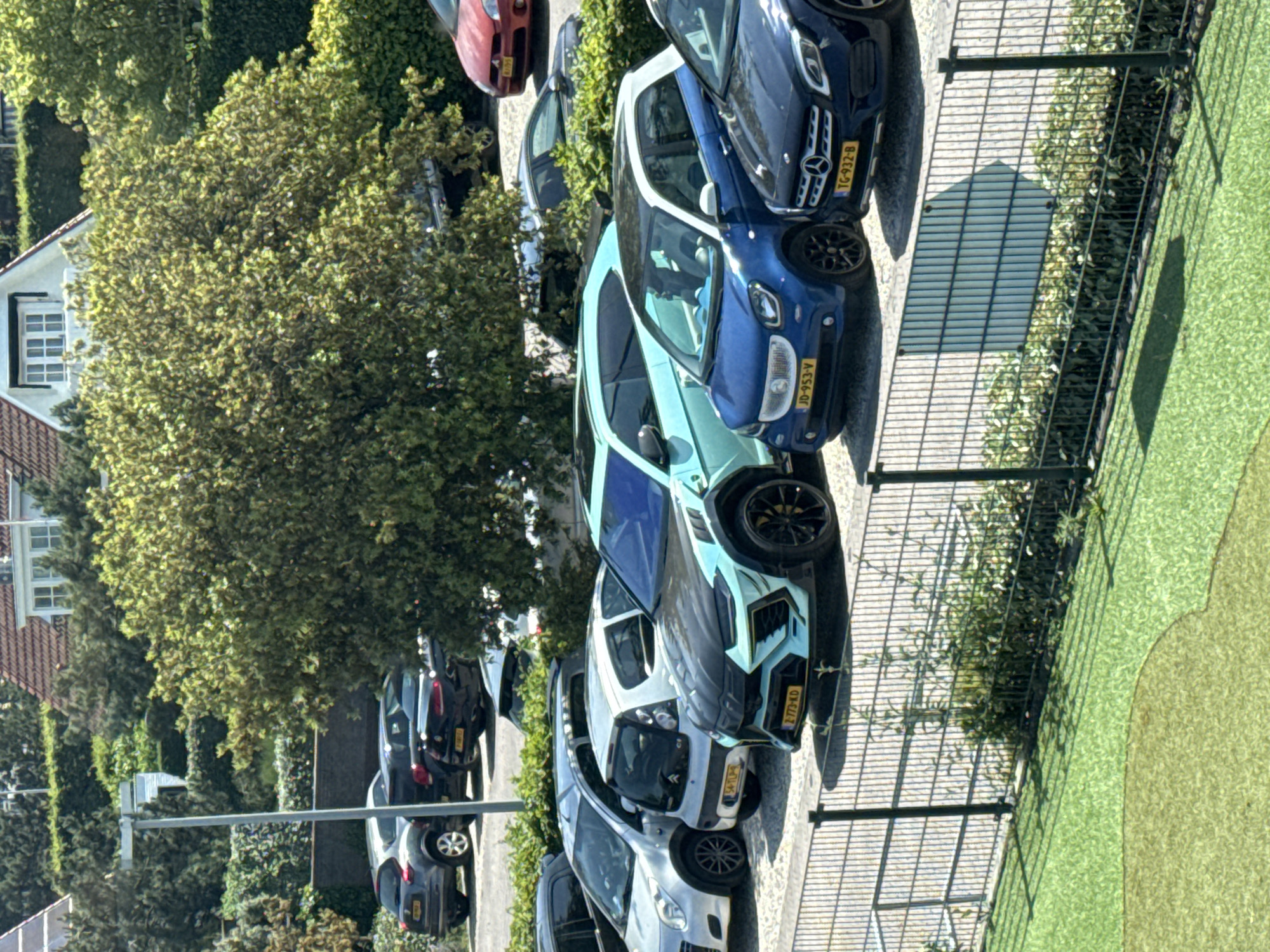 The image captures a street scene in Noordwijk, Netherlands. Multiple cars, including a distinctive light blue and black vehicle with the license plate Z-773-KD, are parked along the street. The parked cars are adjacent to a metal fence and a patch of green grass. A row of buildings and trees are seen along the street on the left.