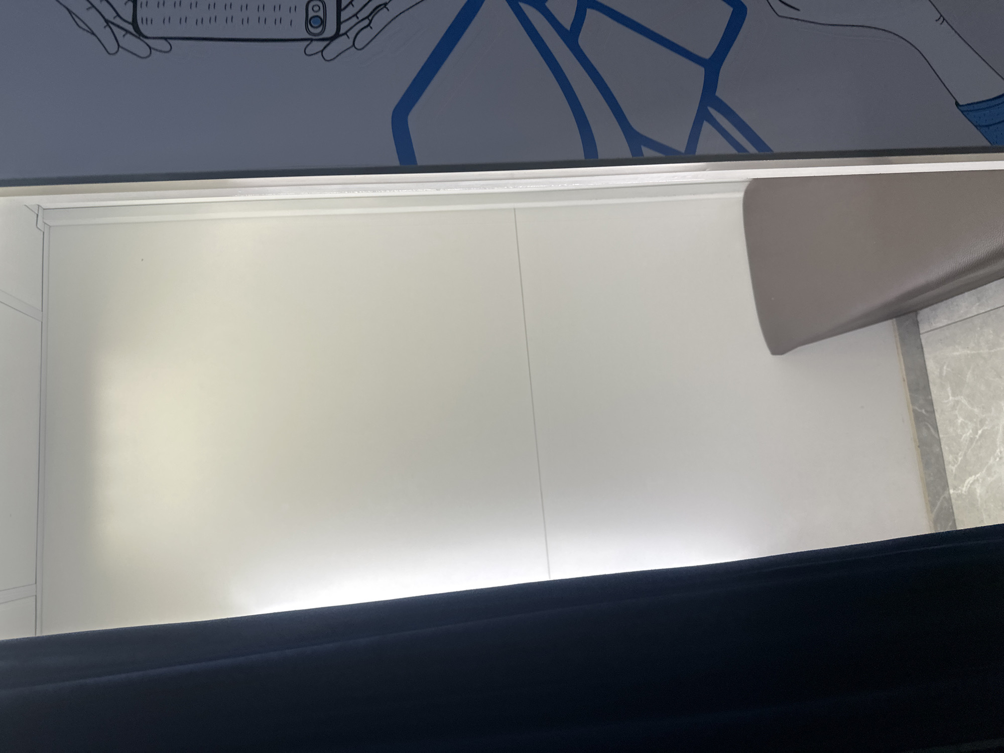 The image shows the interior of a vehicle, possibly a bus or train, in an enclosed space. The upper portion displays a graphic design featuring hands holding a mobile phone and a blue geometric pattern on a white background. Below, there is a clean, white surface, likely an interior wall or panel, with a vertical seam indicating two adjacent sections. To the right, a beige seat or cushion is visible, partially protruding into the space. A dark, presumably black, fabric or material covers the lower portion of the frame. The location, Yerevan, Armenia, is not discernible from the visual information provided.