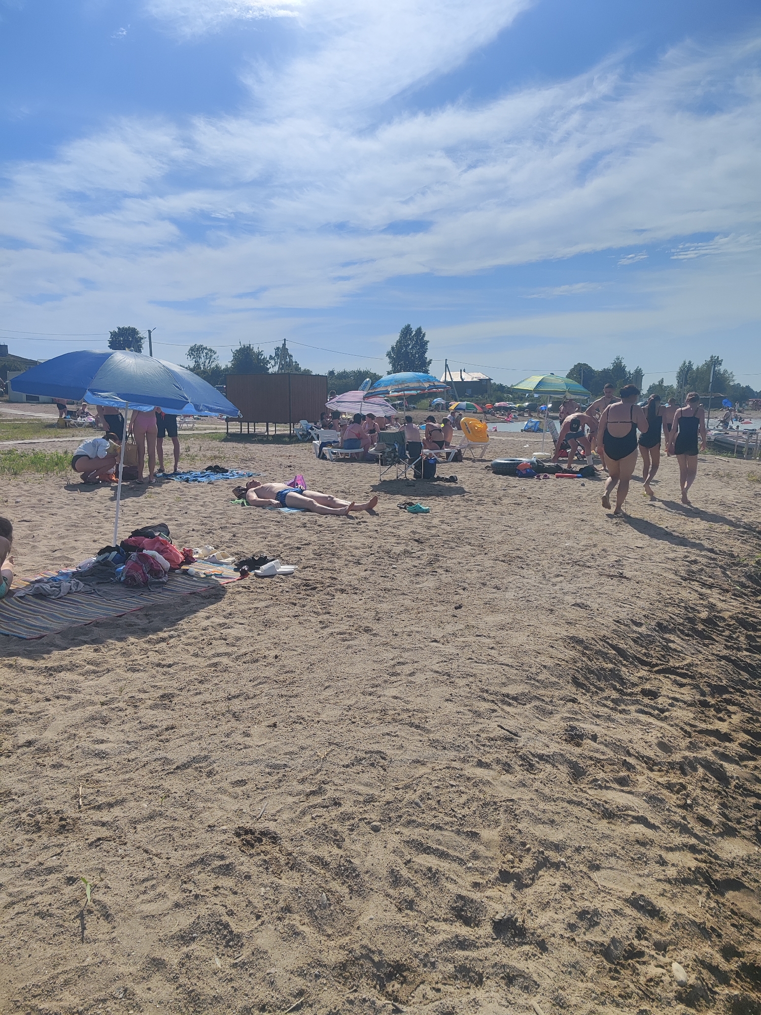 The image depicts a crowded beach scene on a sunny day, with a clear blue sky and scattered white clouds. The beach is sandy, with several individuals engaged in various activities. In the foreground, a man lies on his back on a towel, appearing to be sunbathing. Nearby, a striped mat is spread out with several items of clothing and footwear.
Further back, a group of people are gathered under and around a large blue umbrella. One person is crouching, attending to something on the ground. Several other umbrellas are visible across the beach, providing shade for sunbathers and groups of people relaxing.
In the middle ground, a collection of beach toys, including a yellow inflatable ring and a black inner tube, are scattered on the sand. Several people are walking towards the water, which is visible in the background. The environment appears to be a recreational area with trees and buildings in the distance. The location is identified as the city of Rovnoe, Russia.