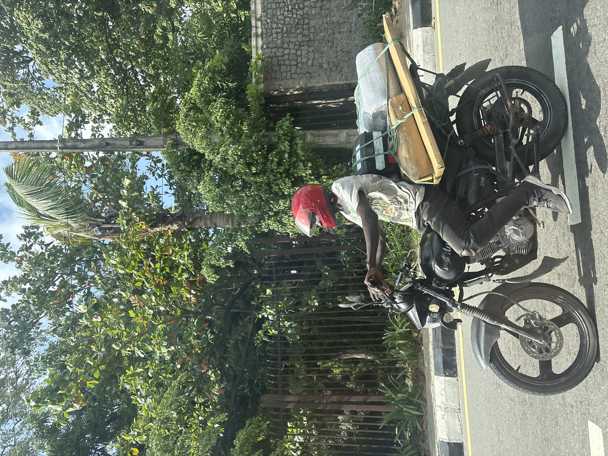 A man wearing a red helmet rides a motorcycle along a road. The motorcycle is heavily loaded with items secured to the back, including a white wrapped bundle and wooden planks. The man is wearing a white t-shirt and dark pants. The background includes lush green trees and a stone wall. The location is Ikoyi, Nigeria.