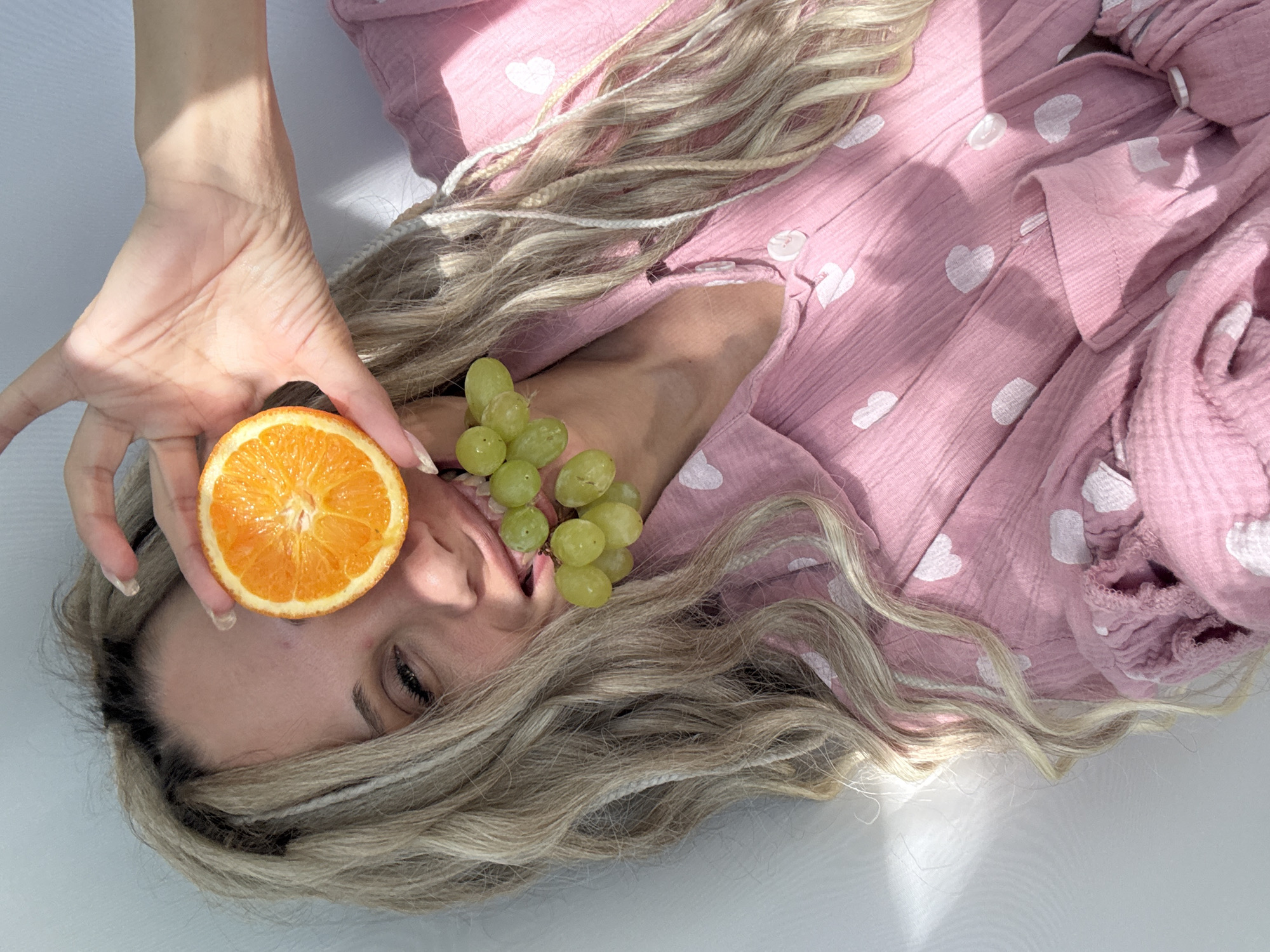 A person lies on their back wearing a pink garment adorned with white hearts. The person's head is tilted, and their light-colored hair with braided sections spreads out around them. A half-cut orange is held near their forehead by a hand with clear polished nails. A cluster of green grapes is near their mouth. The surface beneath appears to be a light grey or white. The location is not discernible from this image.