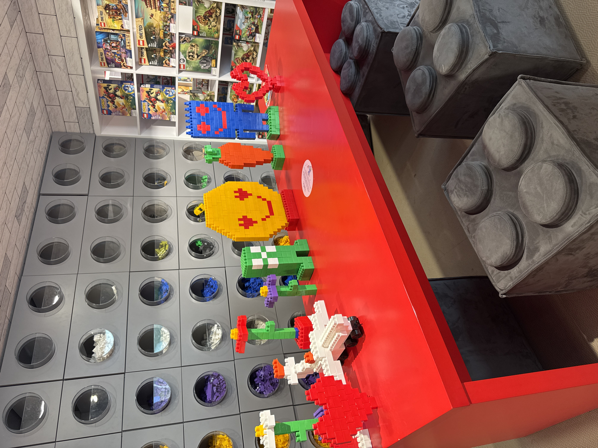 This is a photograph of a display of Lego-themed items in what appears to be a retail or exhibition space. The primary focus is a bright red table laden with various Lego creations. Among these are a large yellow smiley face, a green Creeper-like figure, an orange carrot, a blue robot, a red heart, a white airplane, and several colorful Lego flowers.
Behind the table, a wall is lined with circular displays, each containing an assortment of colored Lego bricks. The wall is constructed of modular grey panels, and above it, a textured ceiling resembling brickwork is visible. To the right of the Lego brick wall, a white shelving unit is stocked with various Lego sets, featuring colorful boxes with images of characters and scenes, including what appear to be boxes for "Sonic the Hedgehog" and "One Piece" themed sets, as well as dinosaur-themed sets.
In the foreground, two large, grey ottoman-like seats, designed to resemble oversized Lego bricks with circular protrusions, are positioned in front of the red table. The floor is carpeted with a neutral-colored, patterned material. The scene is well-lit, suggesting an indoor environment. There are no people visible in the image. The location is identified as Yekaterinburg, Russia.