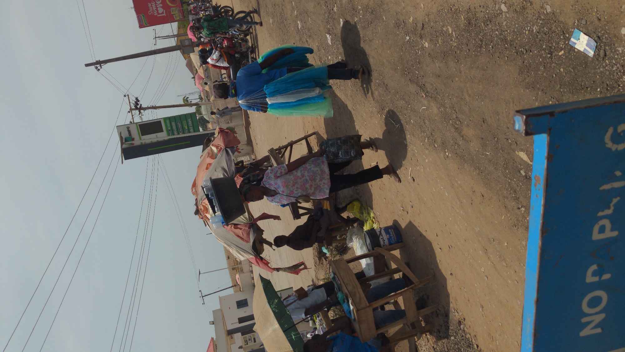 This media file captures a bustling roadside scene, likely a market or commercial area, under a bright, clear sky, indicating midday or early afternoon. The ground is unpaved, consisting of dirt and gravel, with visible litter.
Several people are engaged in daily activities. In the foreground, a person wearing a floral shirt and dark pants stands near a makeshift wooden stall or table laden with items, including a black basin and water bottles. Further back, another individual in a bright blue shirt carries a large, colorful bundle of light blue, white, and green fabric or umbrellas on their back, suggesting they are a street vendor. Various other people are visible in the background, including some seated on wooden structures and others operating or riding motorcycles.
The setting is characterized by utilitarian infrastructure, with multiple wooden utility poles and a dense network of electrical wires crisscrossing the upper portion of the frame. In the background, a tall, vertical sign with green and white elements is prominent, displaying "MRPO" at its top and likely indicating a business or fuel station. Above this, a red billboard is partially visible, bearing the text "Show some LOVE." To the far right in the foreground, a blue structure, possibly part of a stall or container, shows partially obscured text that reads "NO P LY." (likely part of "NO POLICY" or similar). A blue bucket on the ground further displays "Top-Lux Emulsion."
The scene depicts everyday street commerce and public life in what appears to be a vibrant, informal market environment, consistent with a location like Ado, Nigeria. Distinct shadows are cast by the people and objects, affirming the sunny conditions.