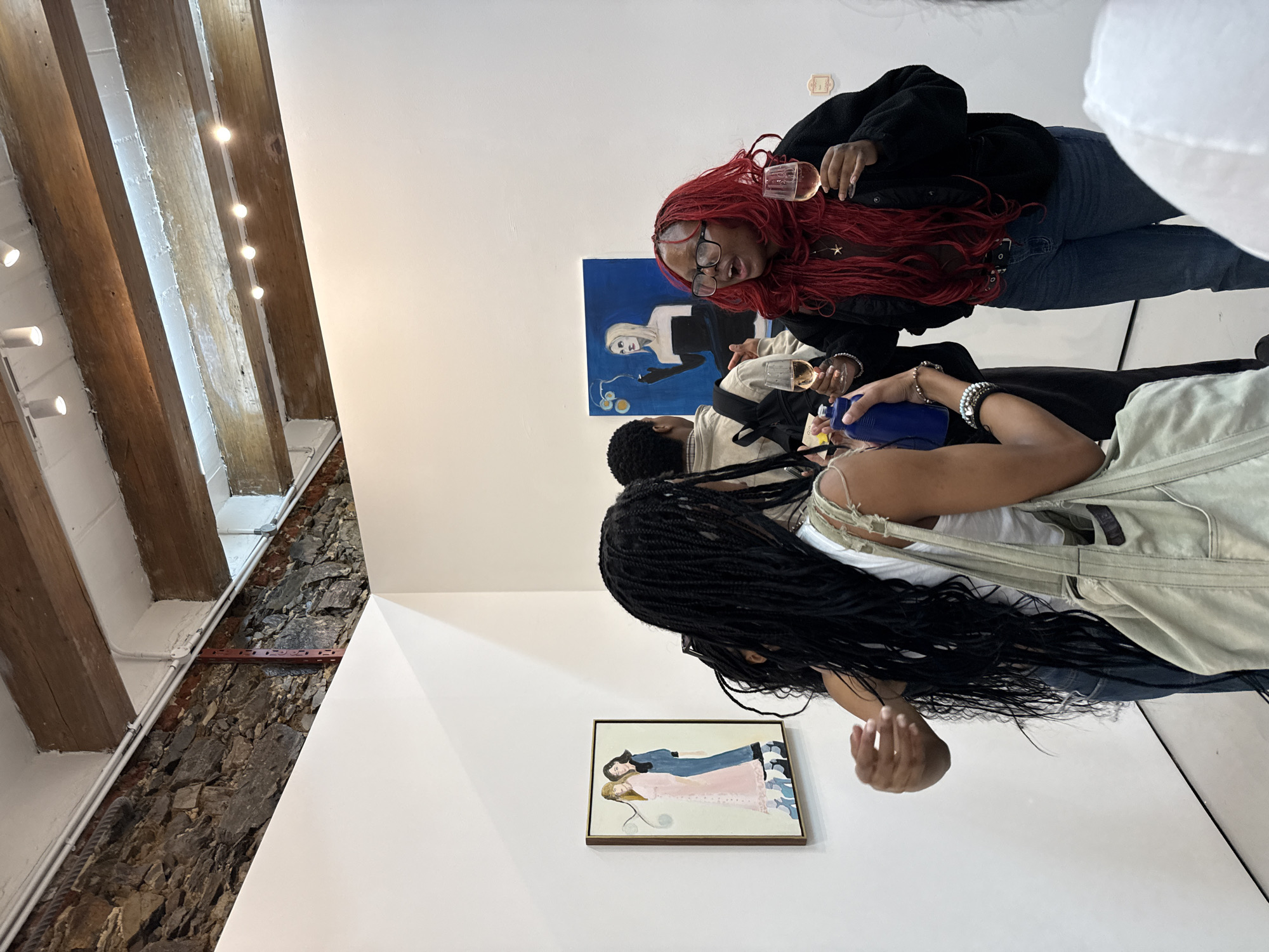 The image shows a group of people in an indoor space, possibly an art gallery, with a visible brick and beam ceiling. Three individuals are partially visible; one woman with long red hair and glasses holds a wine glass. Adjacent to her is another person. The woman in the foreground has long black braids and is wearing overalls. Two framed artworks adorn the walls. The location is Unknown, Unknown.