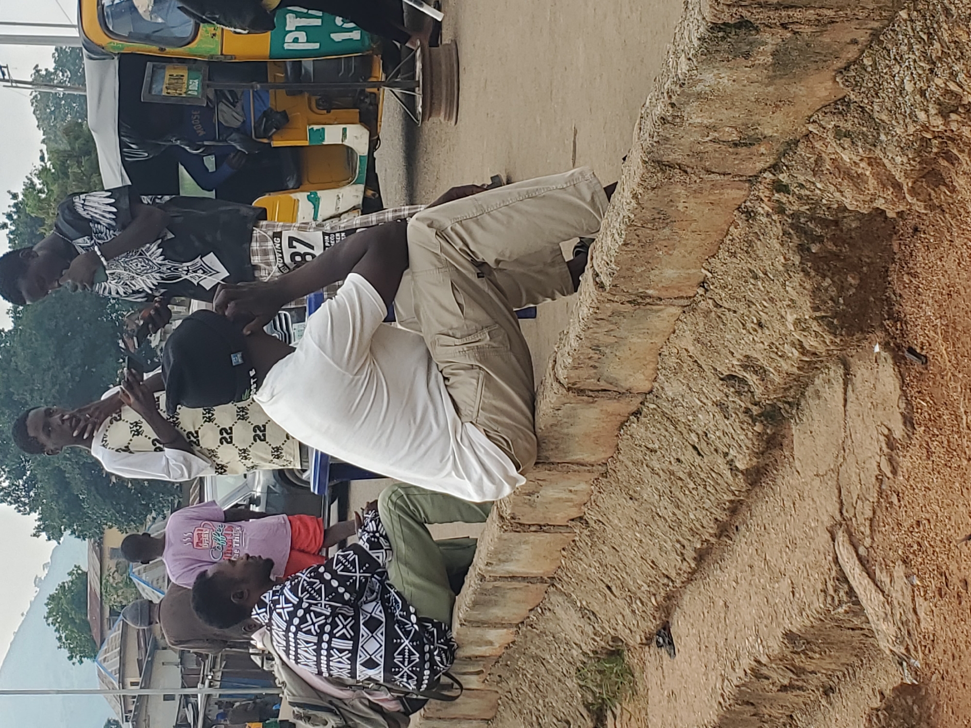 In Gwong, Nigeria, a group of young men are seen in an outdoor setting, likely a street or market area. In the foreground, two men are seated on a concrete barrier. The man on the left, wearing a black and white patterned shirt and green pants, is looking to his left. The man on the right, in a white t-shirt and khaki pants, is looking forward with his hand on his chin. To their right, a yellow and green tricycle, commonly used as a taxi, is parked. Another young man in a black t-shirt with a white design is standing near the tricycle, holding a mobile phone. In the background, several other individuals are visible, including a person wearing a pink t-shirt with text and graphics, and another in a white shirt with a patterned design. Buildings and trees form the backdrop of the scene, suggesting an urban or semi-urban environment.
