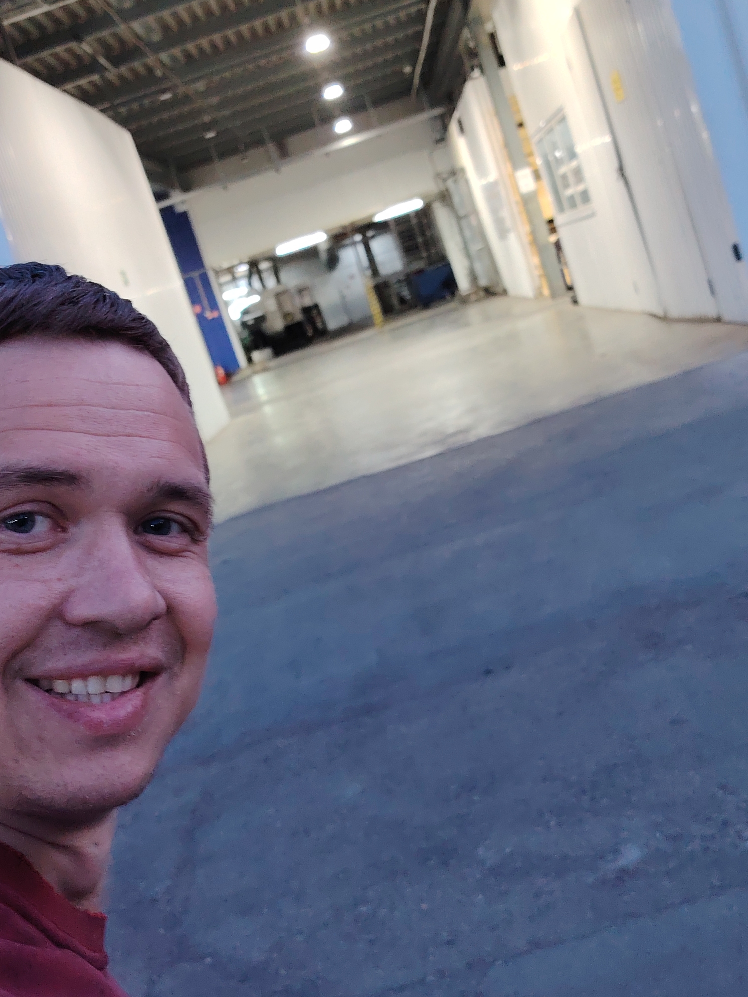 The image is a selfie of a man with short dark hair and light skin, wearing a red shirt. He is smiling. The background is an industrial or commercial space, possibly the interior of a ship or a large warehouse. The space appears to be brightly lit with overhead lights. The walls are white or light-colored. There is equipment or machinery visible in the distance. The floor is gray concrete. The location in the city of Noviy, Russia is undeterminable.