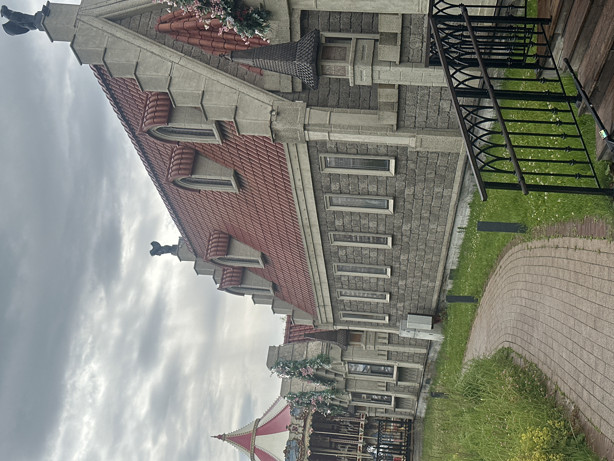 The image shows an outdoor scene with a building that has a unique architectural style, featuring a red tiled roof, stone facade, and decorative elements. A dark metal staircase with intricate railings is visible on the right side of the building, leading down to a paved pathway and a grassy area. There is a hint of a carousel with red and white striped canopy in the lower left corner, suggesting it is part of an amusement park or a similar attraction. The sky is overcast, indicating a cloudy day. The building's design, particularly the gargoyle-like figure on the roof and the detailed stonework, gives it a somewhat whimsical or fantastical appearance. The overall impression is one of a creatively designed building in a park-like setting, possibly in Moscow, Russia, given the location context. No people are visible in the image. The time of day is indeterminate due to the overcast sky.