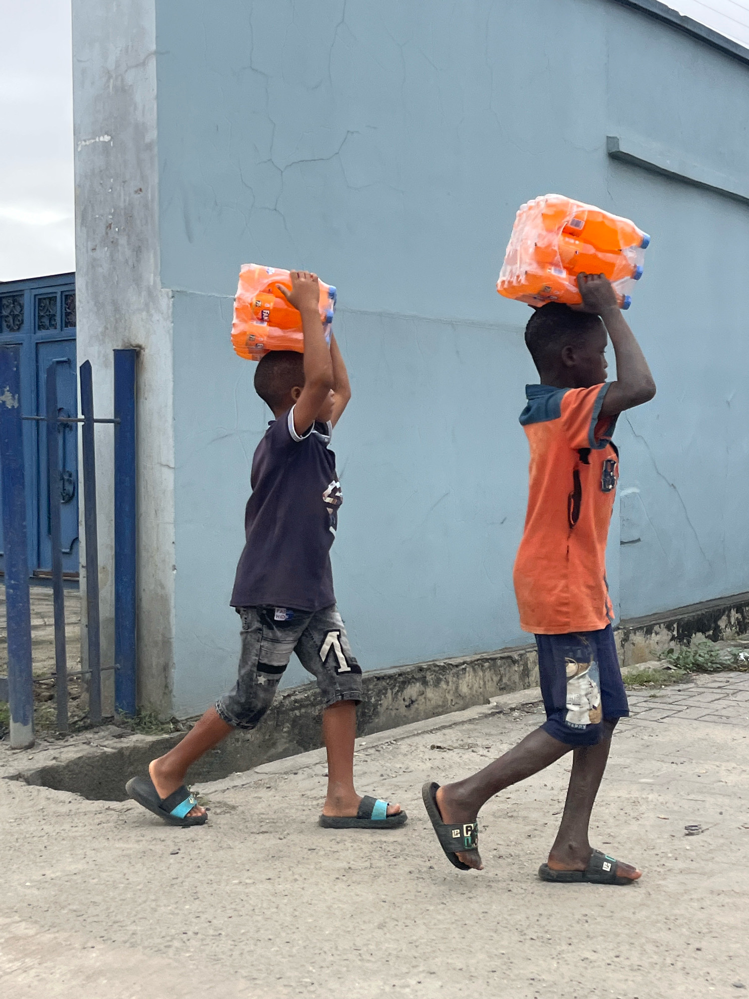 Two young boys are walking along a street in Ado, Nigeria, each carrying a multipack of orange drinks on their heads. The boy on the left wears a dark t-shirt and patterned shorts, and the boy on the right wears an orange t-shirt with dark blue accents and dark shorts. They are walking on a paved surface with a slight incline. Behind them, a light blue building with some visible cracks stands next to a blue metal fence and gate. The sky is overcast.