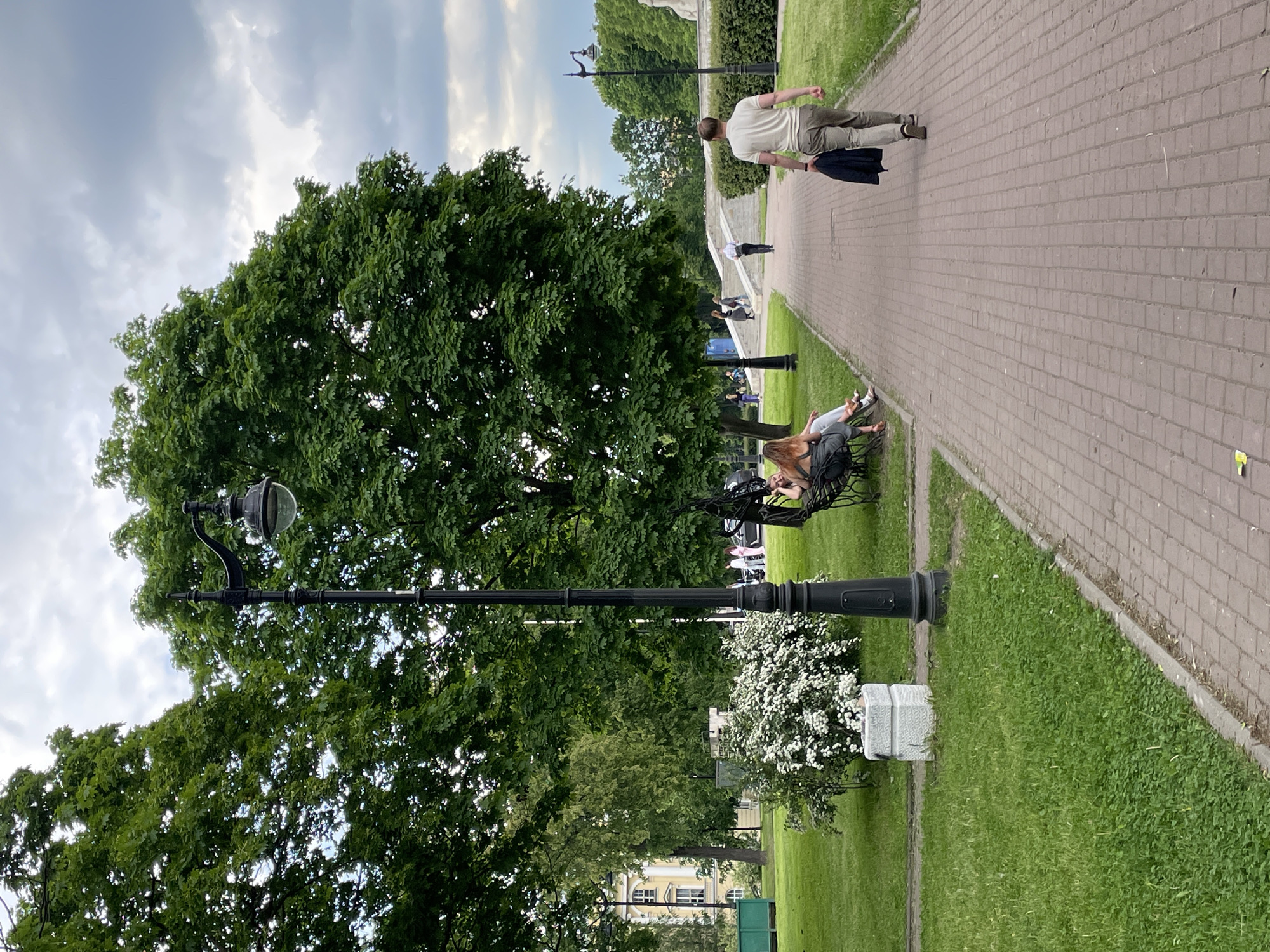 The image captures a tranquil scene in a public park or garden, identified as being in Saint Petersburg, Russia. The perspective is slightly elevated, looking down a wide paved brick path that extends diagonally from the foreground towards the background. Green grass and well-maintained lawns border the path on both sides.
On the left side of the frame, a large, mature tree with dense green foliage dominates a significant portion of the view, extending towards a cloudy sky. A tall, black ornamental street lamp with a globe light fixture stands in the mid-ground on the left.
Several people are present, engaged in leisurely activities. On the right side of the paved path, closer to the foreground, two women are seated on a dark park bench. Further down the path, a man in a light-colored shirt and trousers walks away from the viewer, holding a dark jacket. Other indistinct figures can be seen walking further in the distance and on a set of stairs.
Notable details include patches of white flowering bushes near the base of the street lamp and a white stone monument or pedestal adjacent to it. In the background, partially obscured by trees, a building with yellow walls and white window frames is visible, suggestive of classical European architecture. The sky is largely overcast with light clouds, indicating daytime, possibly late afternoon, with soft, diffused light. No explicit text is clearly legible on any of the visible objects. The scene conveys a peaceful atmosphere, characteristic of a public park setting.