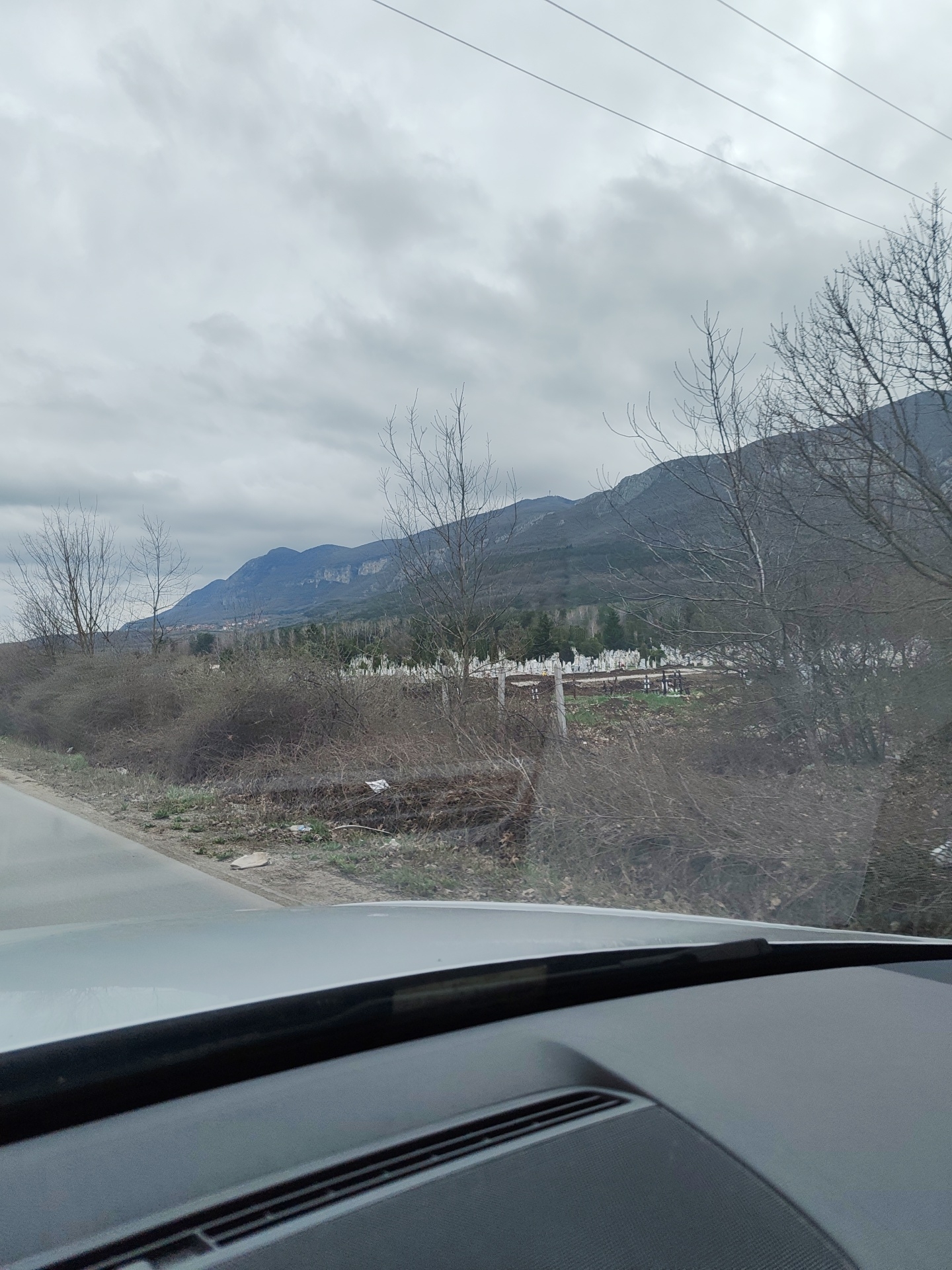 The image shows a view from inside a car, looking out over a landscape. In the distance, there's a mountain range under a cloudy sky. In the midground, there's a cemetery with white headstones. In the foreground, there is roadside vegetation. The photo appears to be taken near the city of Vratsa, Bulgaria, given the specific type of landscape and cemeteries in the area. The camera is positioned behind the dashboard of a car, parts of which are visible.