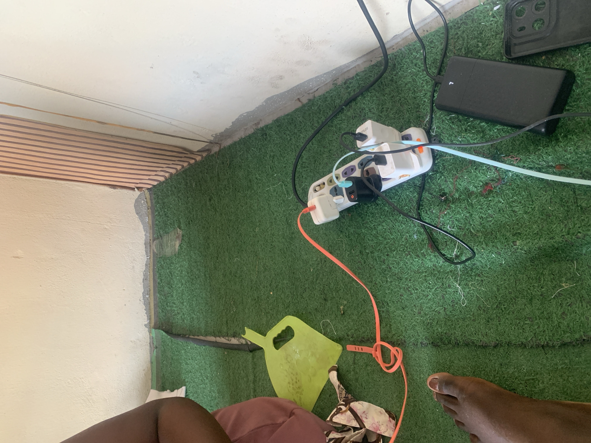 The image depicts an indoor scene with a predominantly green, artificial turf floor. A white wall, showing signs of wear and damage near the base, forms the backdrop. To the right, a vertical element with vertical brown lines suggests a textured wall or screen. In the foreground, a person's left foot is visible, positioned on the turf.
Several electrical devices are arranged on the turf. A white power strip is plugged into an outlet, with multiple adapters and charging cables connected. One orange cable is looped and tied with a plastic tie. A black rectangular power bank is also connected and appears to be charging. Next to the power bank, a black phone case is partially visible. A bright yellow plastic item, possibly a small shovel or scoop, lies on the turf to the left of the power strip. A patterned fabric item is also present.
The overall impression is a functional, albeit somewhat cluttered, setup for charging electronic devices within a basic interior space. The lighting suggests it is daytime. The location is Jauro Yinu, Nigeria.