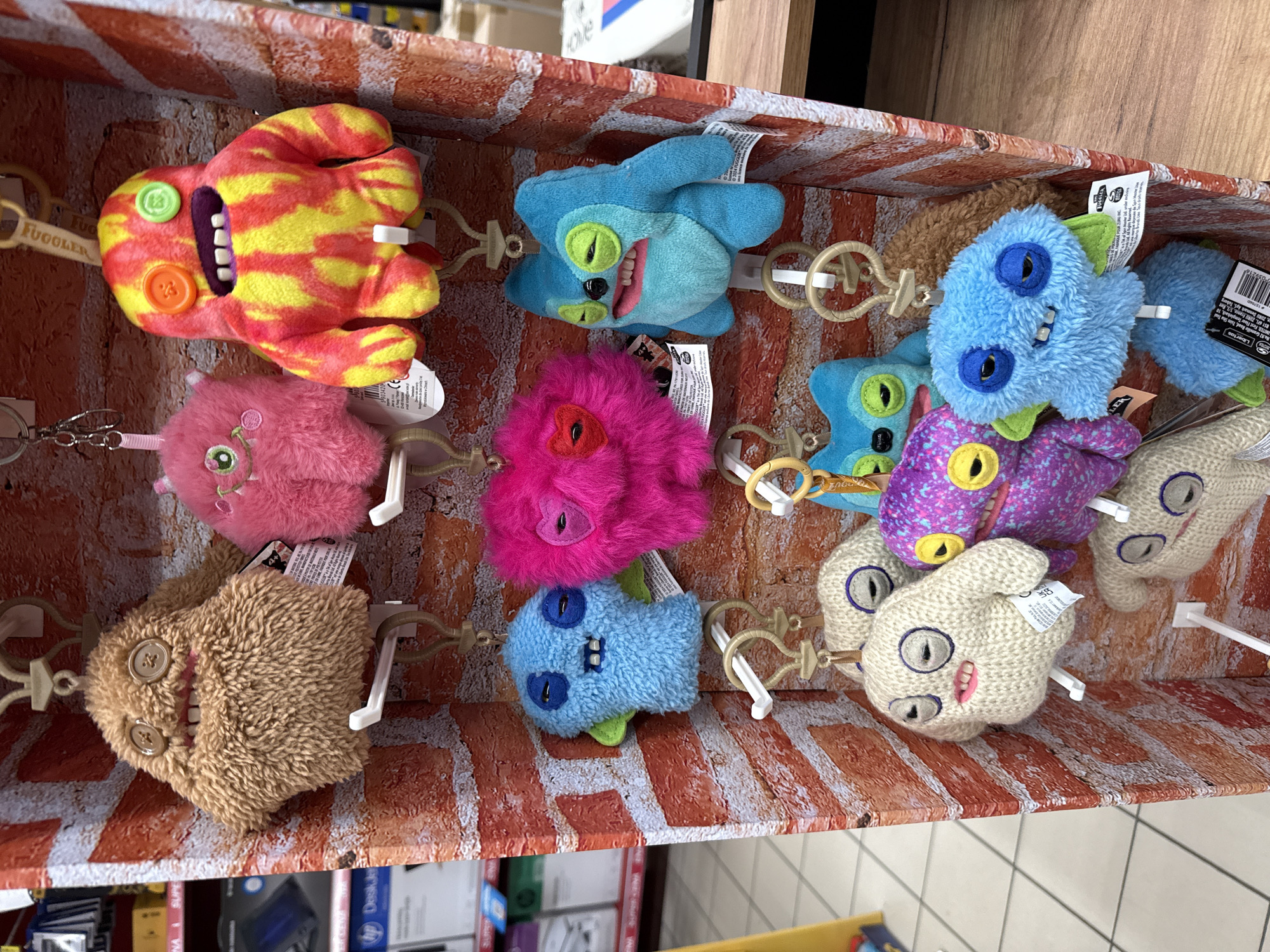 The image displays a retail display of plush monster-themed keychains. The keychains are arranged in rows on a brick-patterned cardboard structure. There are at least nine visible keychains, varying in color, texture, and facial features, with most featuring large eyes and open mouths with teeth. The background suggests a retail environment with shelves and products visible on the left side. This display is located in Wrocław, Poland, specifically within the Empressi store at ul. Legnicka 58.
