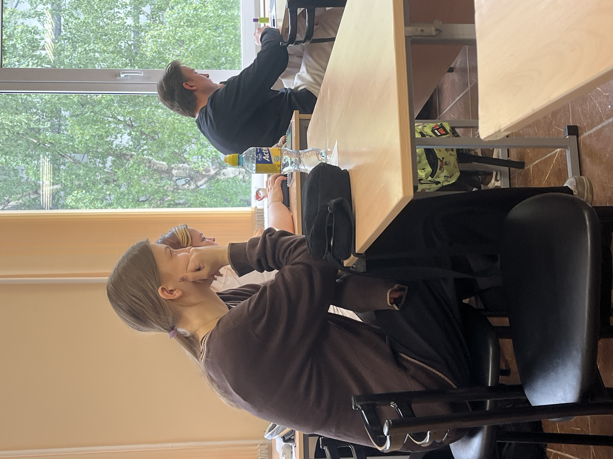 A classroom scene shows two individuals seated at desks. The person in the foreground is a young woman with long brown hair tied back in a low ponytail with a purple hair tie. She is wearing a brown zip-up sweatshirt and is resting her chin on her hand, seemingly engaged in a conversation with someone out of frame. The table in front of her has a black soft bag resting on it.
In the background near the window, a young man is seated at another desk, holding an object up to his eye as if viewing something. A bottle of water is on the desk in front of him. A window is visible behind the individuals and shows a view of trees outside, revealing the greenery of an outdoor environment.
Based on the architecture and context, this image appears to be taken in a classroom setting, most likely at the university in Saint Petersburg, Russia.