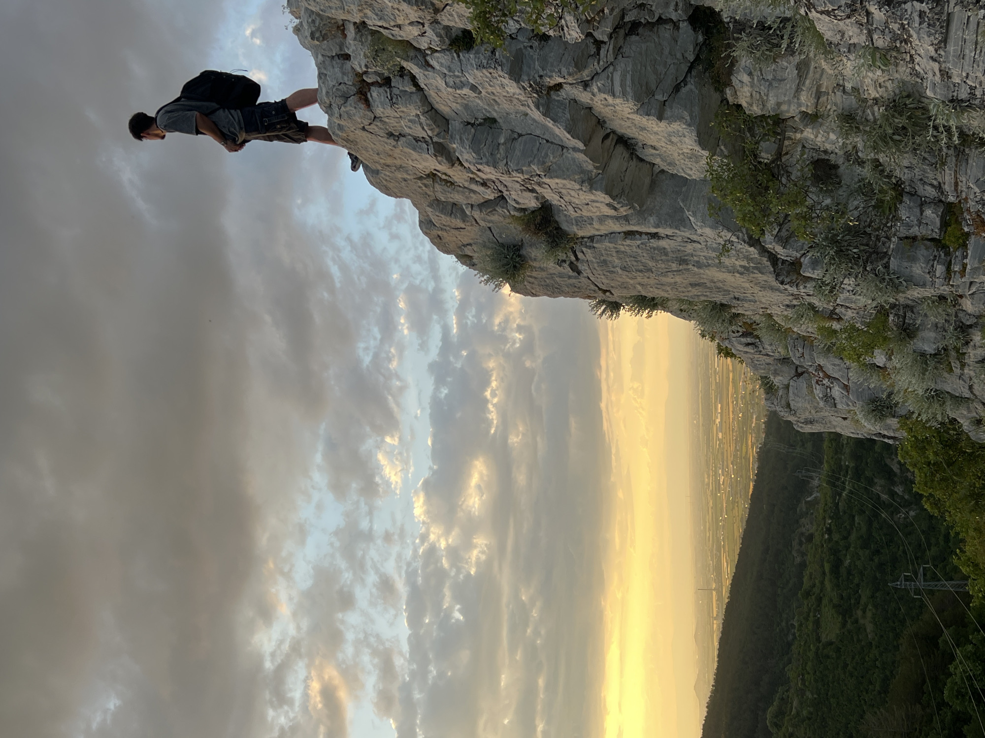 The image shows a person lying prone at the edge of a cliff. The person is wearing a gray shirt, dark shorts, and shoes, and has a black backpack. The cliff is made of gray rock with some vegetation growing on it. Below the cliff, there is a view of a city, which may be Asenovgrad, Bulgaria, with fields and forests visible. The sky is cloudy with a yellowish hue.